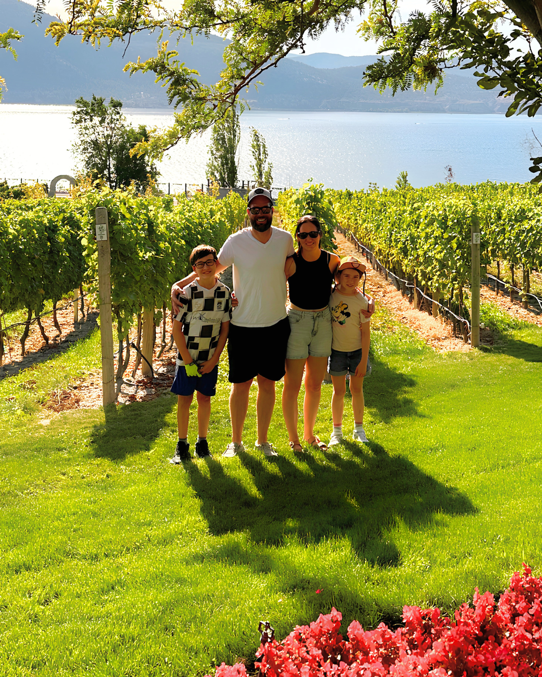 A family of four standing together in a vineyard near a body of water with mountains in the background. They are outdoors on a sunny day, smiling and posing for the photo.