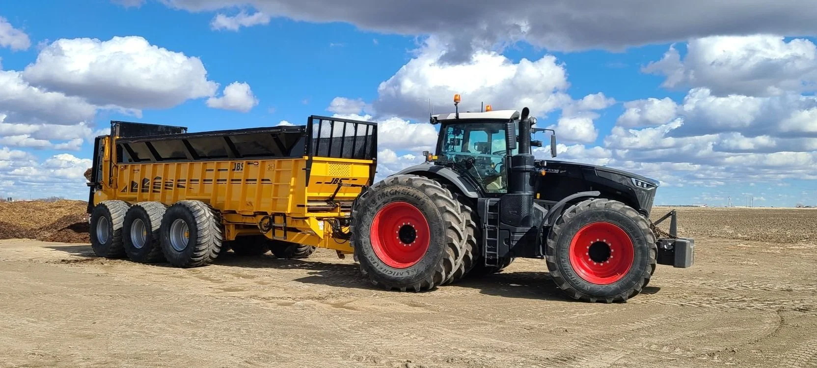 A large Fendt 1050 tractor pulling a yellow trailer on a dirt field, under a partly cloudy sky.