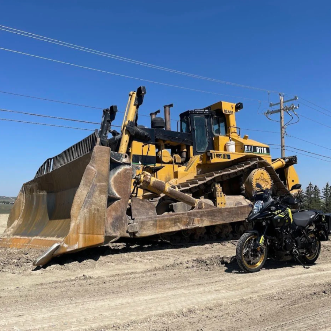 A large yellow Caterpillar bulldozer with a rusted blade parked on a dirt road under a clear blue sky, with a black motorcycle with yellow accents and saddlebags in front of it.