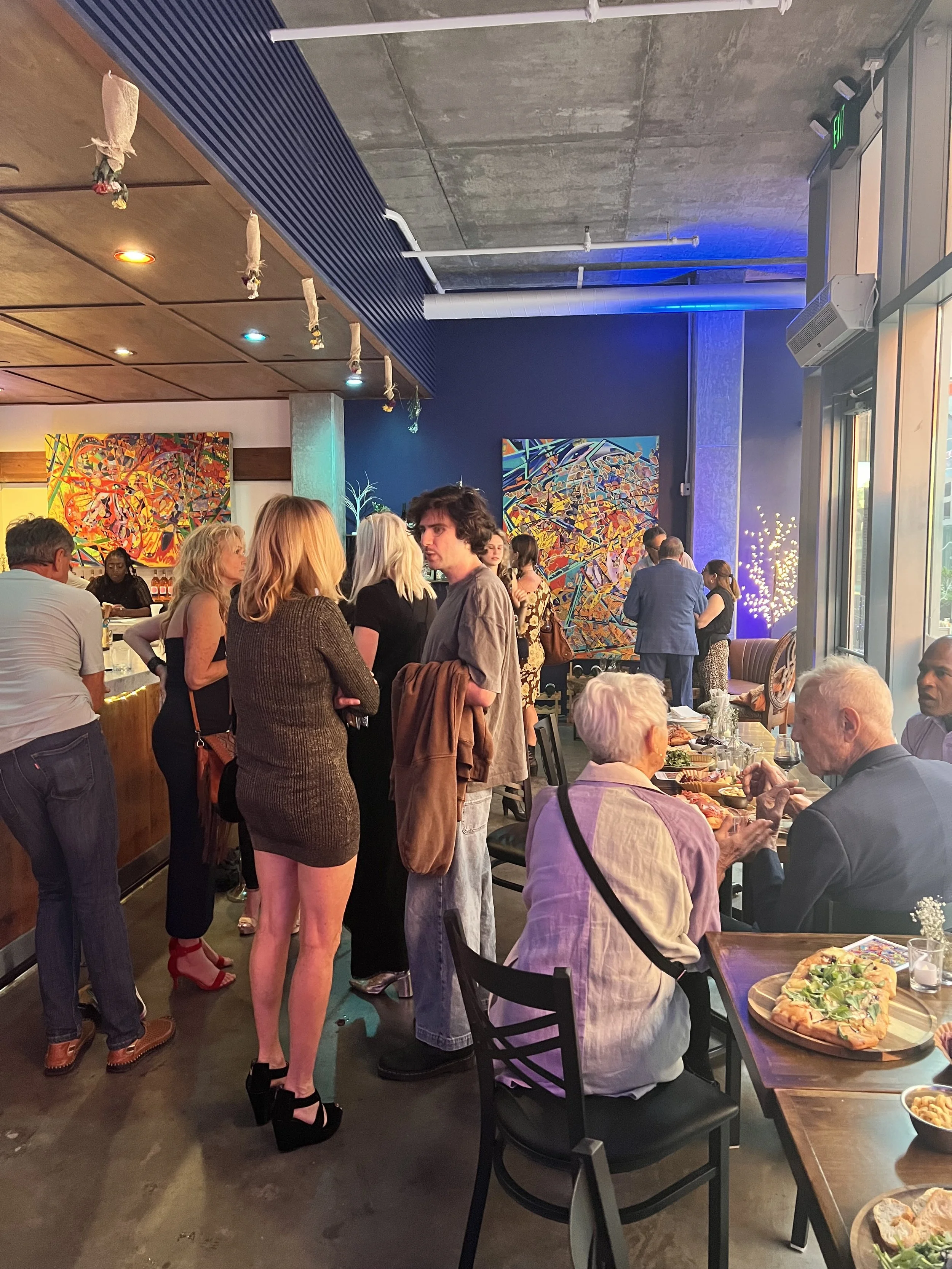 People mingling and dining at a social gathering in an art-filled indoor space, with tables of food and colorful paintings on the walls.