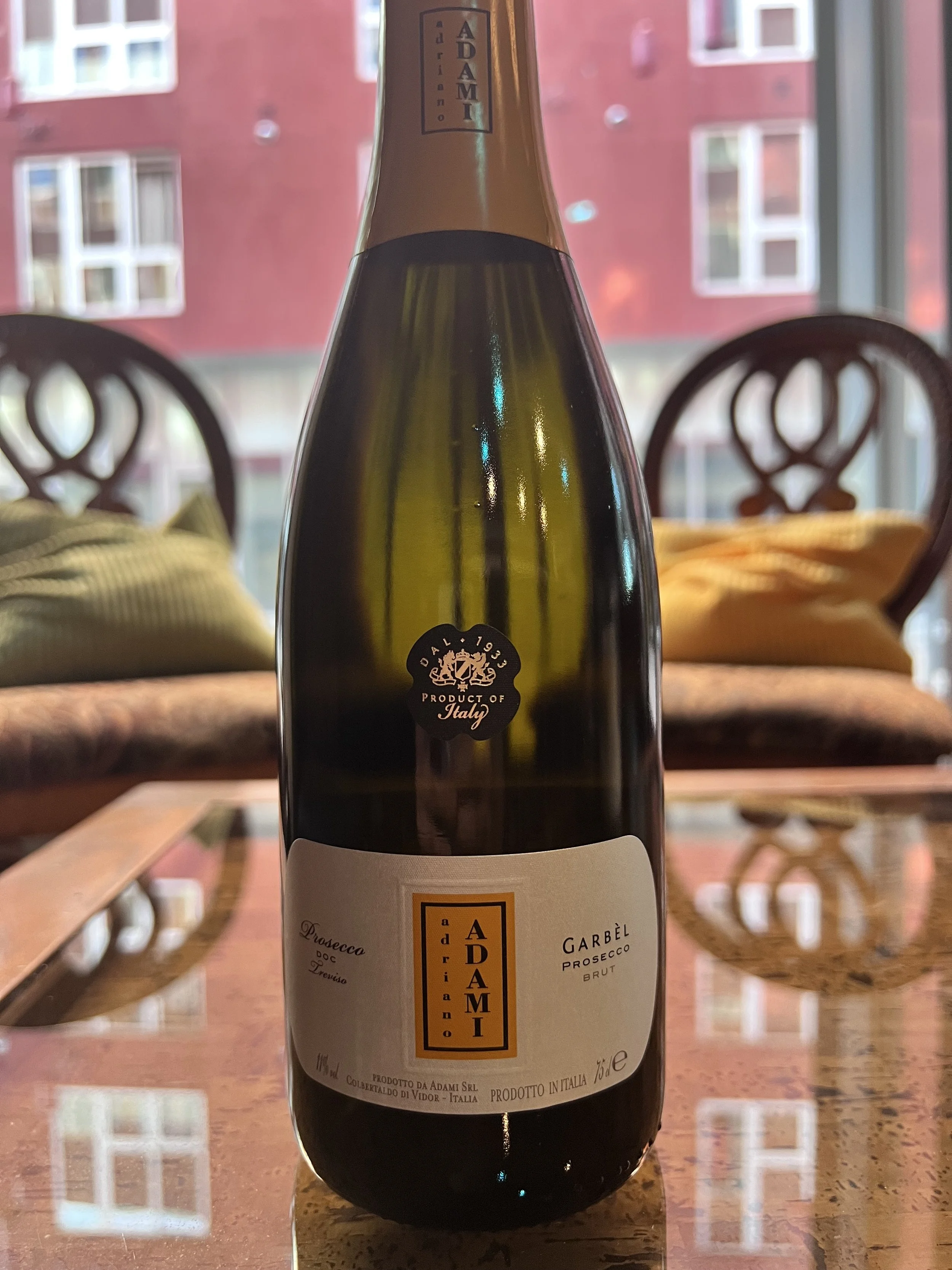 A bottle of Adami Garbel Prosecco Brut placed on a polished wooden table in a room with large windows and blurred city buildings in the background.