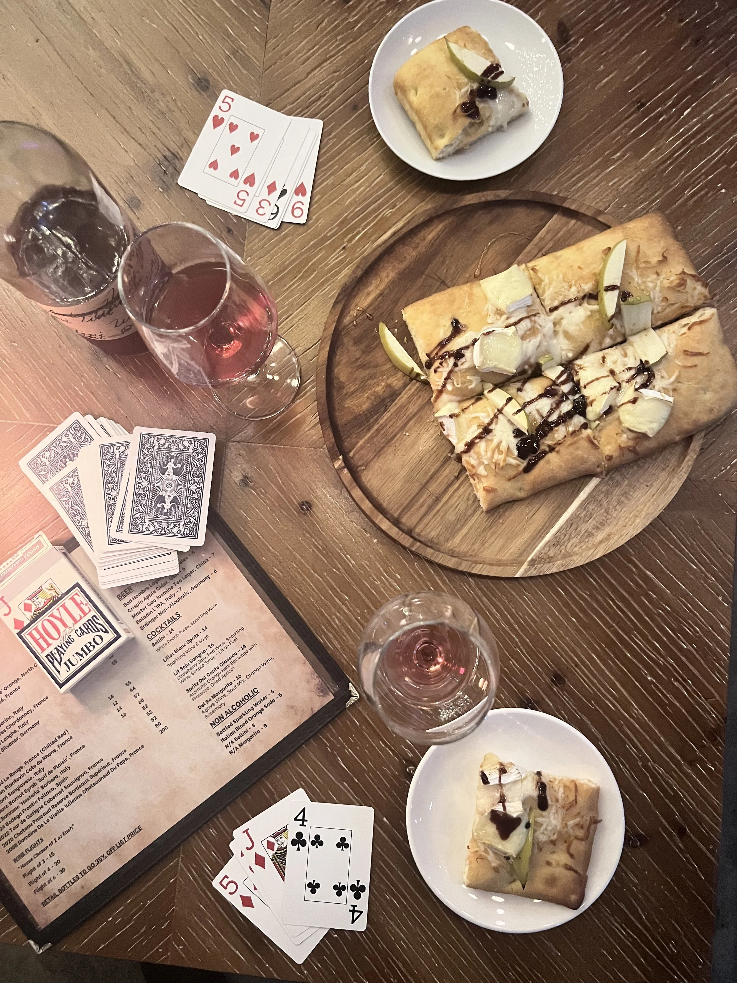 A table with two glasses of pink wine, a deck of playing cards, a menu, two plates with crepes topped with pizza that has apple slices and hot honey , and a round wooden serving board with a larger pizza or flatbread and some additional toppings.
