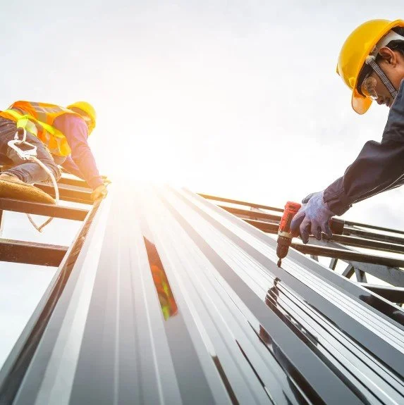 Two construction workers wearing safety helmets and reflective vests work on a metal structure with a screwdriver, with the bright sky in the background.