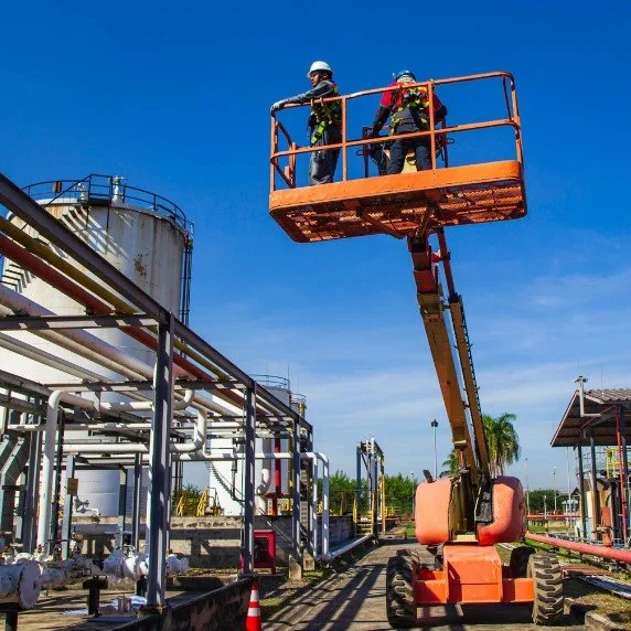 Two workers wearing safety helmets in an elevated work platform at an industrial facility.