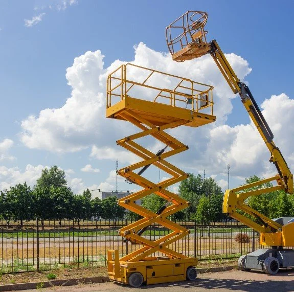A yellow aerial lift with a scissor-style extendable arm and a work platform at the top, positioned outdoors on a sunny day with trees and a fence in the background.