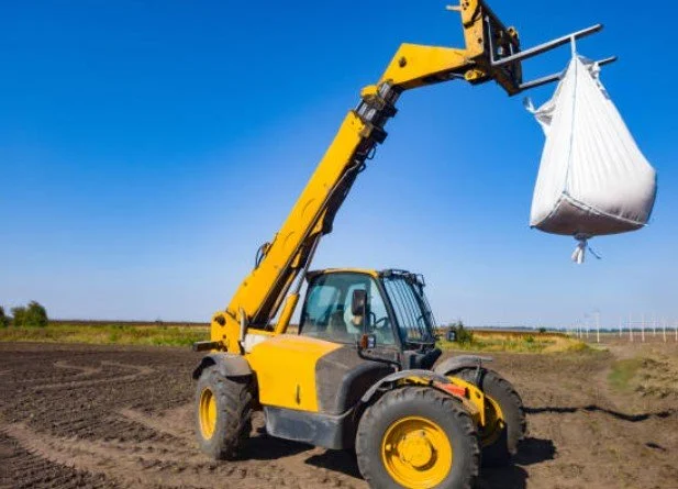 A yellow telescopic forklift lifting a white bag in an open field.