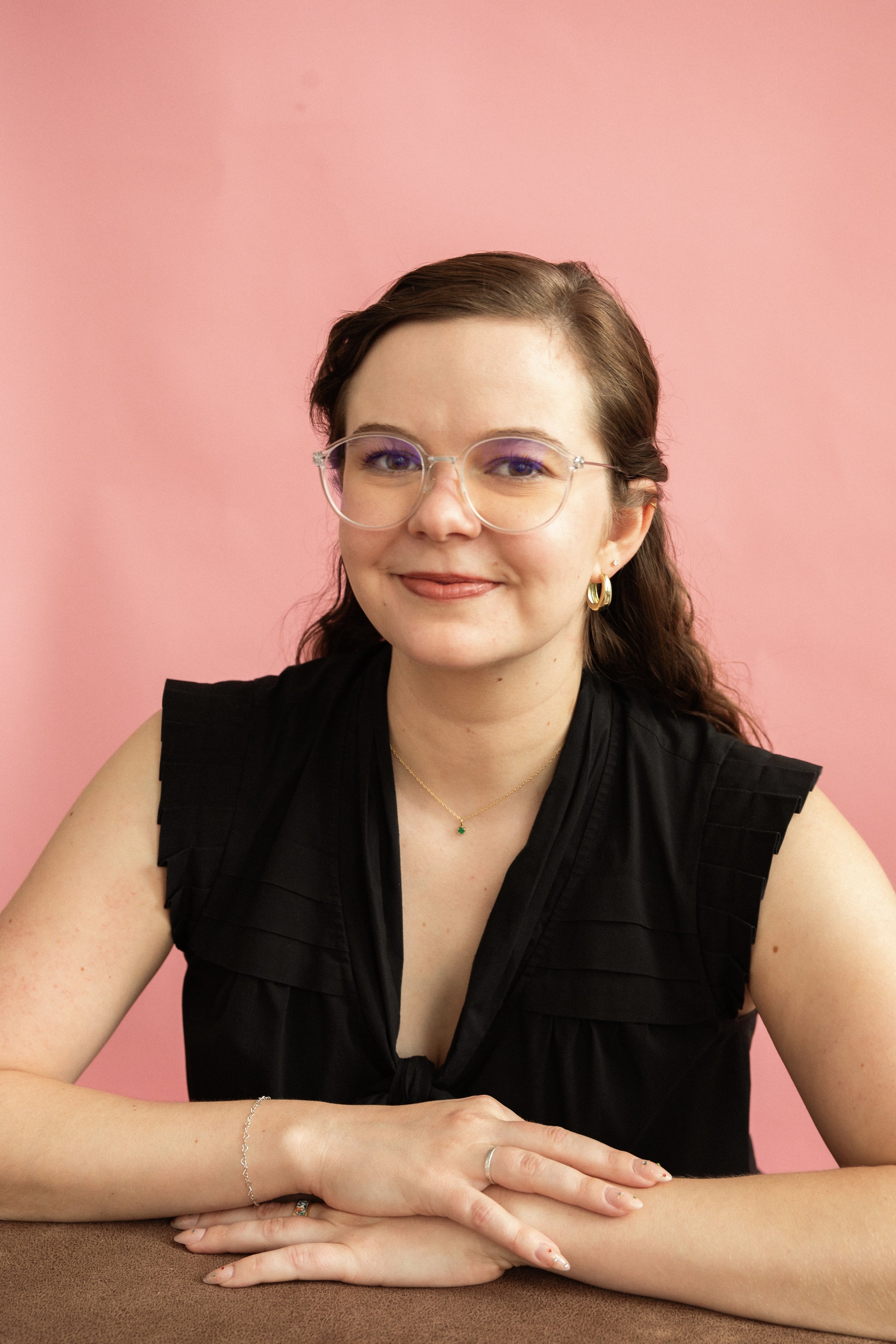 A woman with brown wavy hair, wearing glasses, a black sleeveless top, gold hoop earrings, a gold necklace with a green stone, and a silver bracelet, sitting with her arms on a brown table against a pink background.
