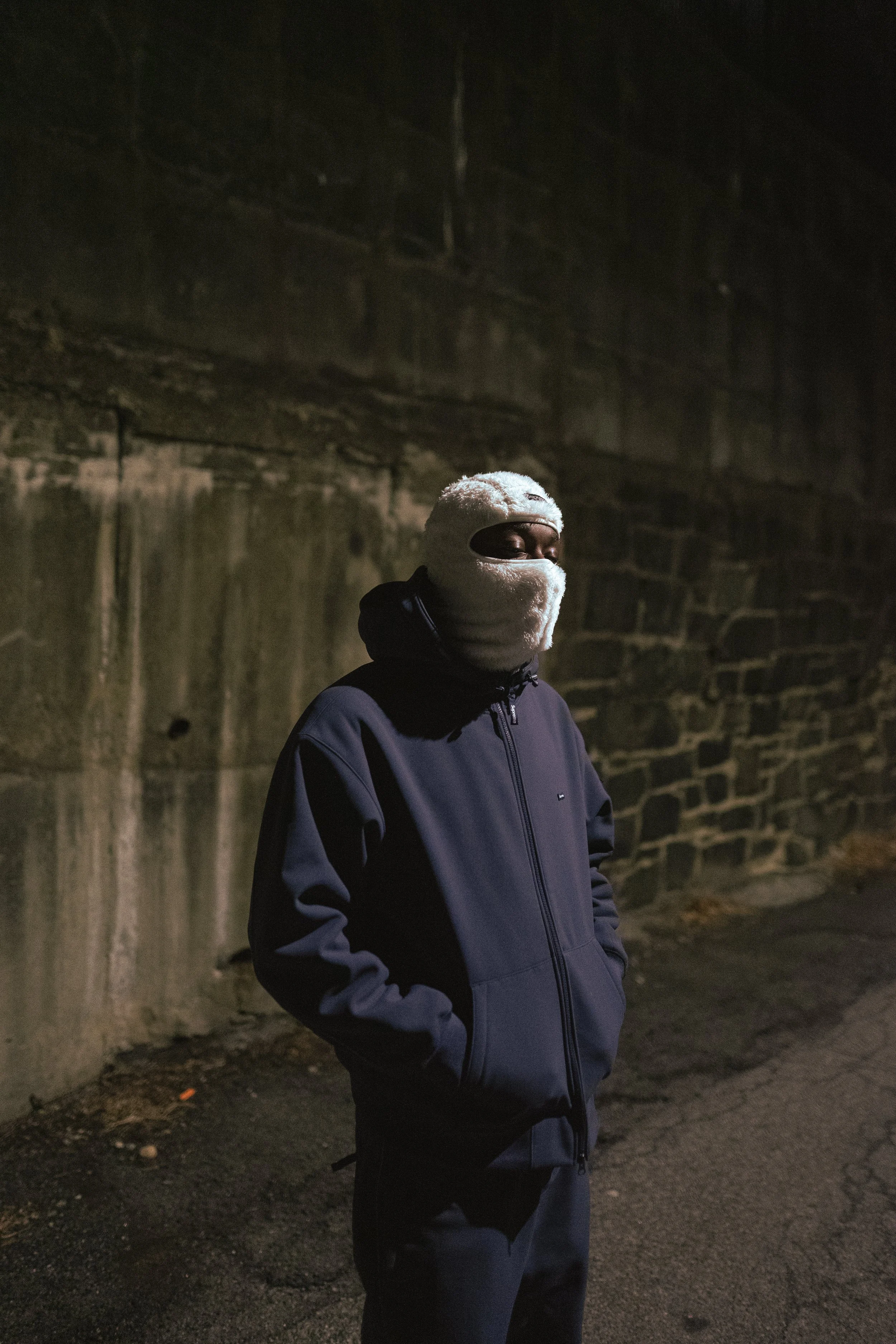 Person wearing a dark jacket and white ski mask standing against a stone wall at night.