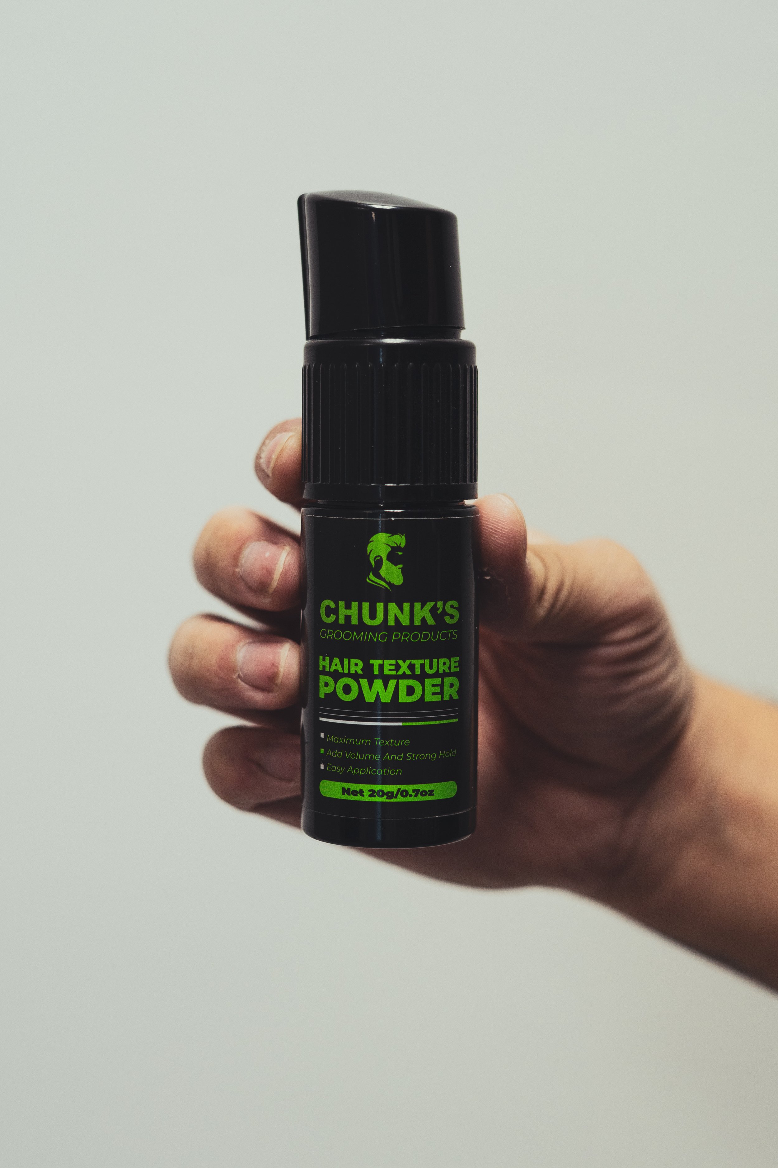 Hand holding a black bottle of Chunk's Hair Texture Powder with green text and logo against a plain background.