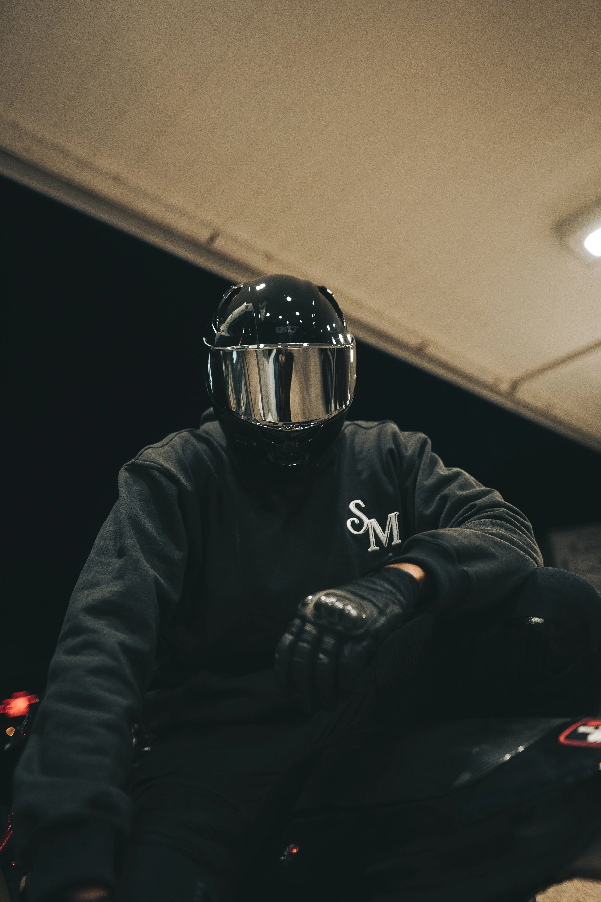 Person wearing a reflective motorcycle helmet, black jacket, and gloves, sitting on a motorcycle indoors.