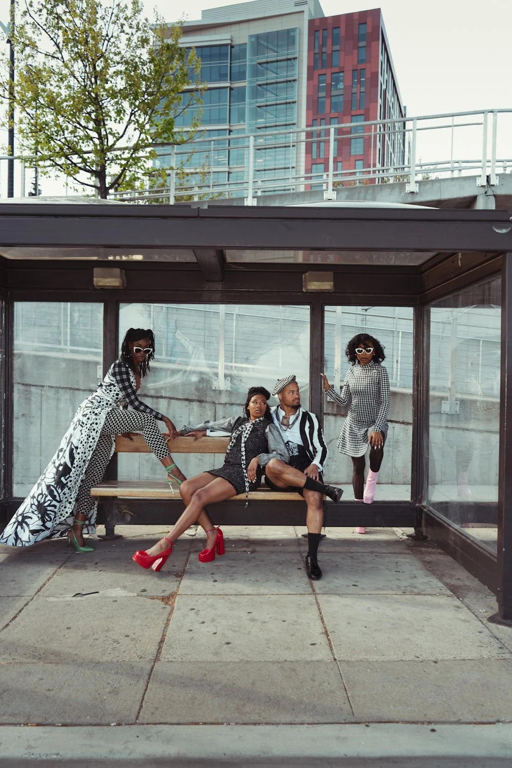 Four stylish people in black and white outfits with colorful shoes posing at a bus stop in an urban setting.