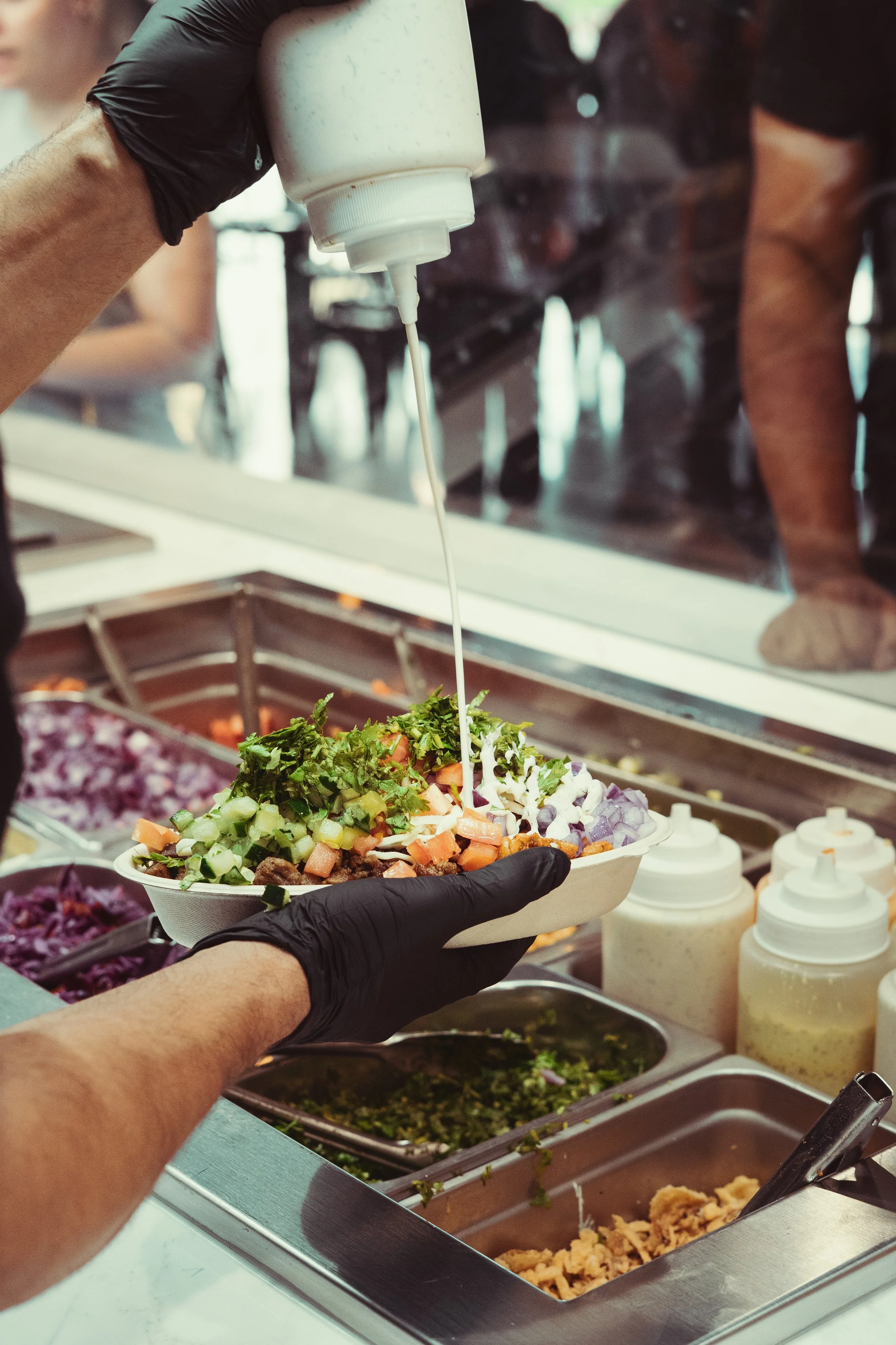 Person with black gloves pouring white dressing on a plate of salad, with vegetables and toppings, in a food service setting.