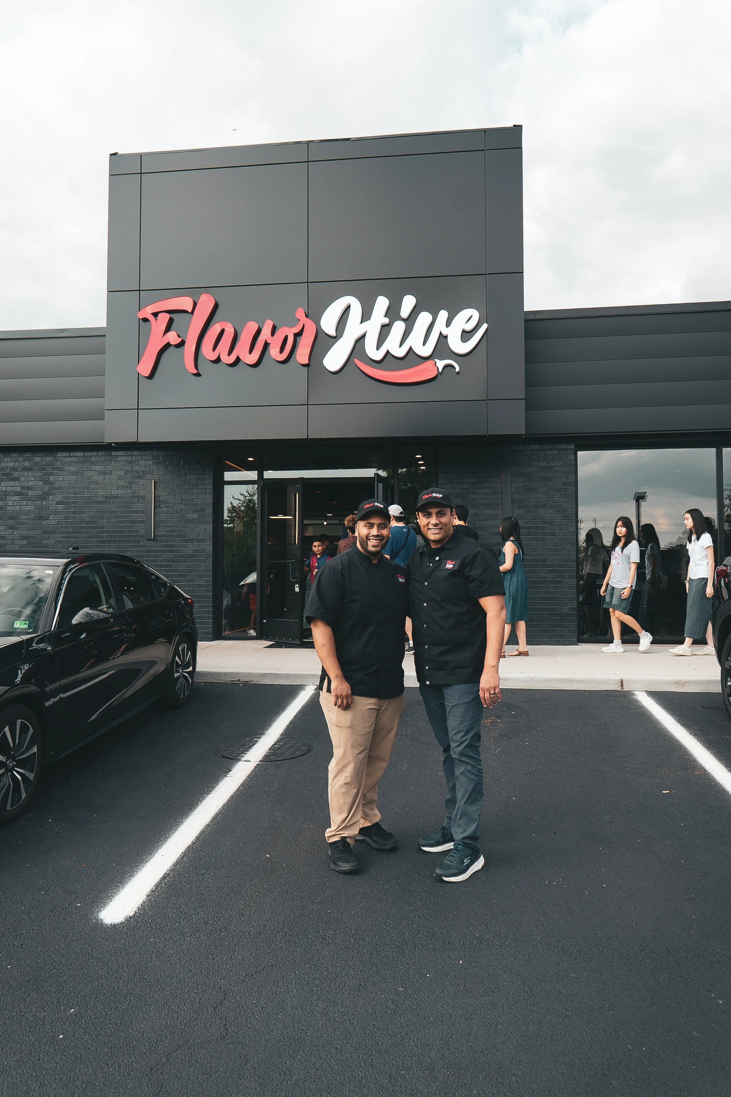 Two employees standing in front of the Flavor Hive restaurant during opening day, smiling and wearing black uniforms, with customers inside and parking spaces visible.