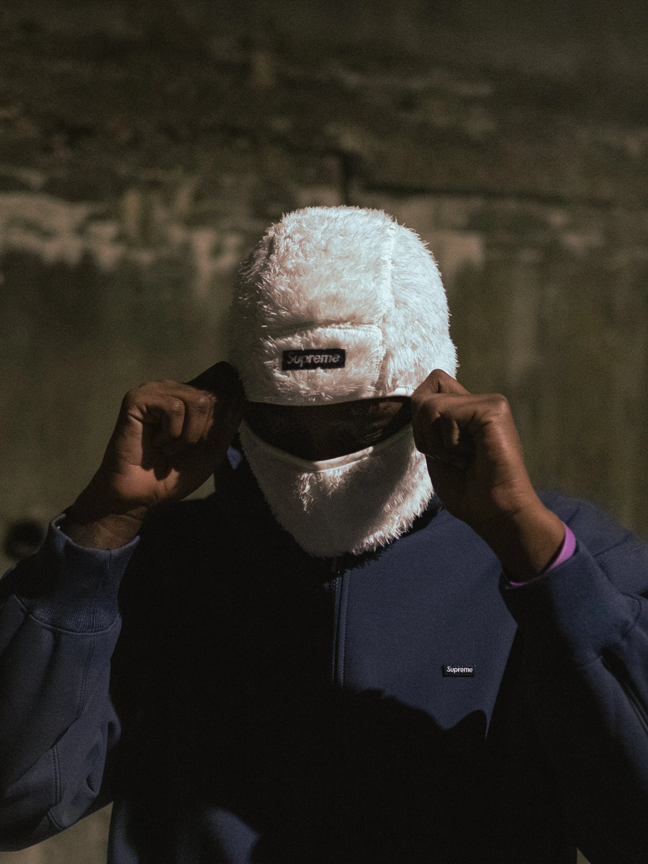 Person wearing a blue jacket and a white fuzzy balaclava with "Supreme" logo in a dimly lit setting.