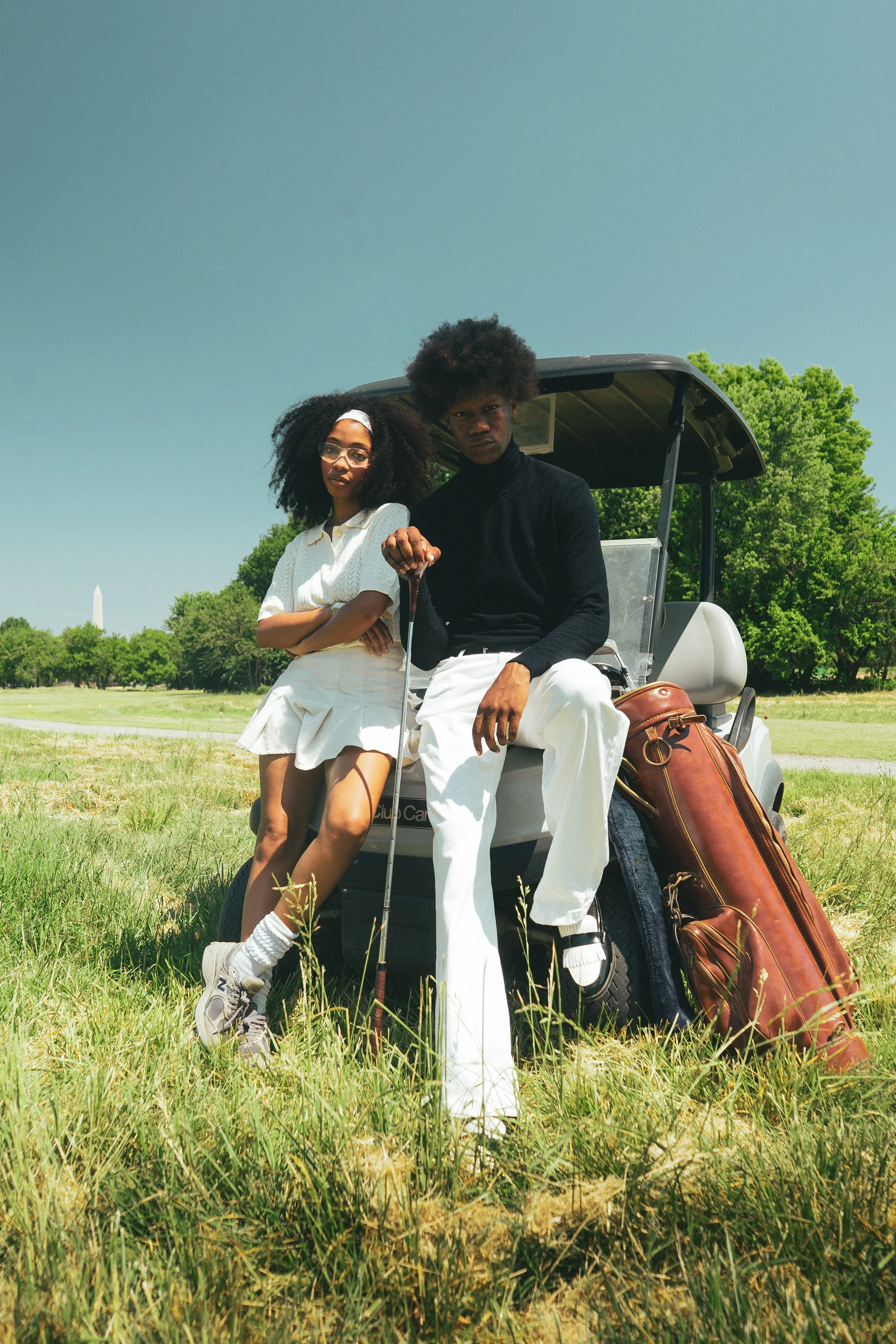 Two young adults, a woman with glasses and curly hair and a man with an afro, sitting on a golf cart on a golf course. The woman wears a white outfit and sneakers, the man wears a black turtleneck and white pants, holding a golf club. Green trees and