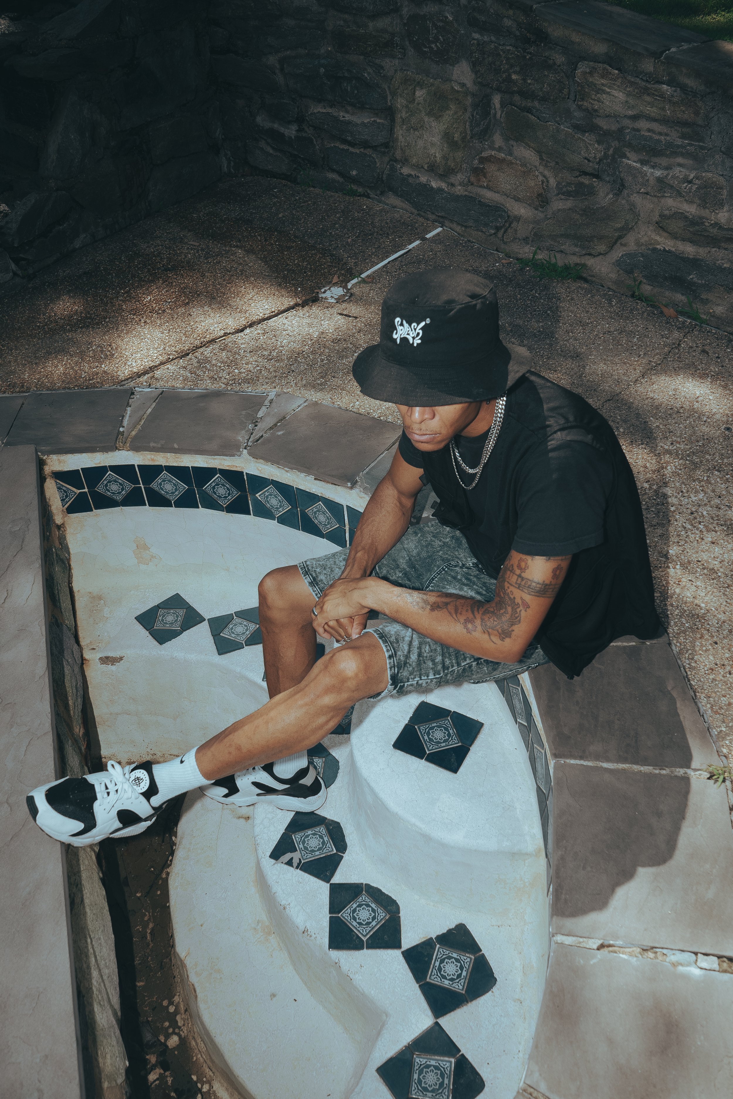 A man wearing a black bucket hat, black t-shirt, and gray shorts sits on the edge of a small circular decorative tiled fountain or pond, with broken and intact tiles, in an outdoor area with stone walls.