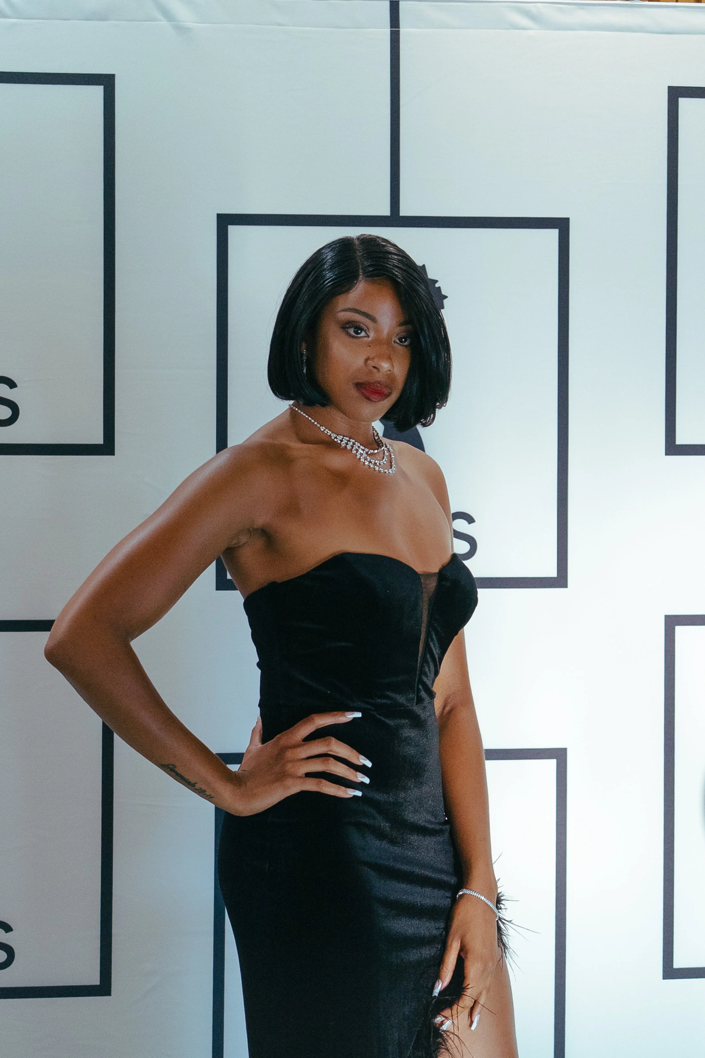 A woman in a strapless black dress with a slit, wearing a diamond necklace and earrings, posing with one hand on her hip against a white background with black framed text.