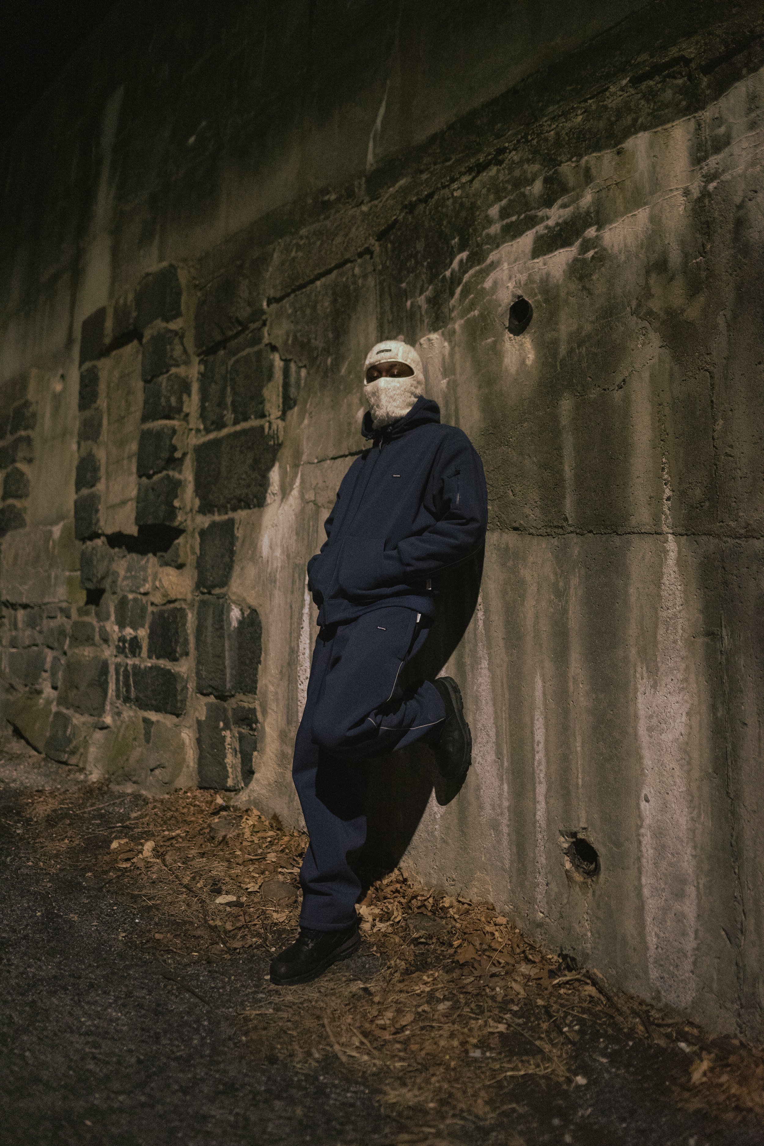 Person in dark clothing and ski mask leaning against a stone wall.