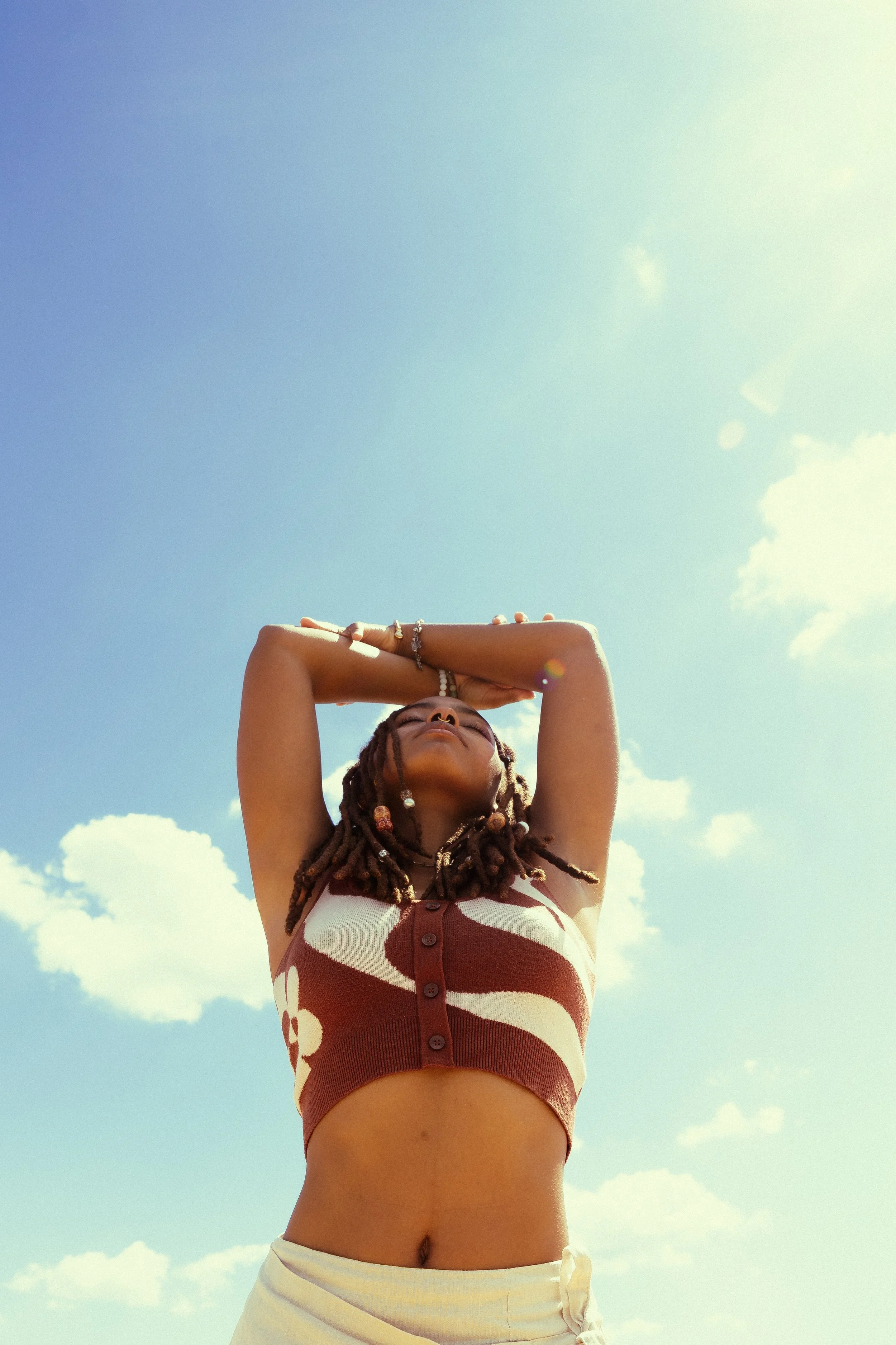 A woman with dreadlocks posing outdoors under a blue sky with scattered clouds, wearing a patterned sleeveless top and cream-colored shorts.