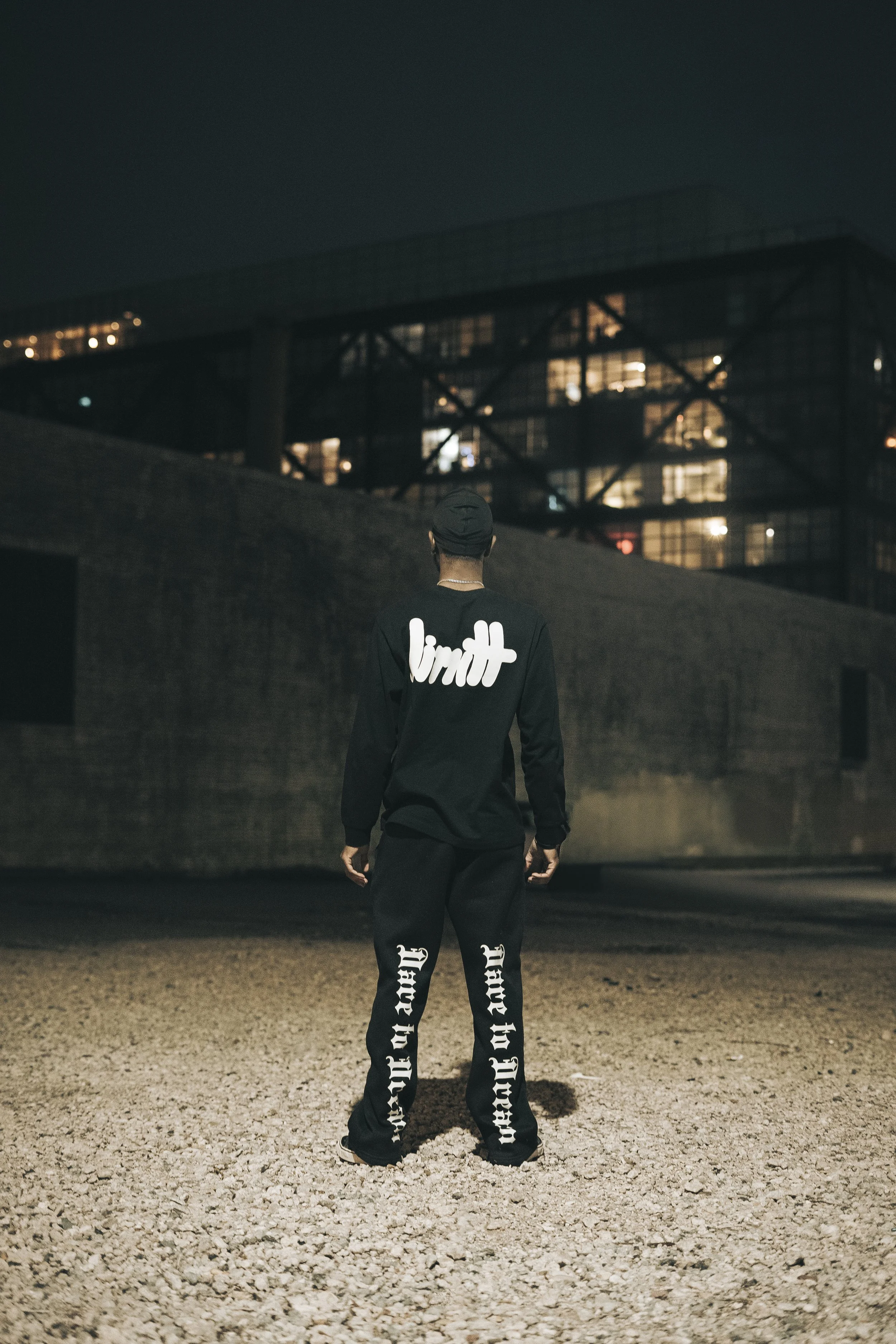 Person in black clothing standing outside at night, facing a lit industrial building.