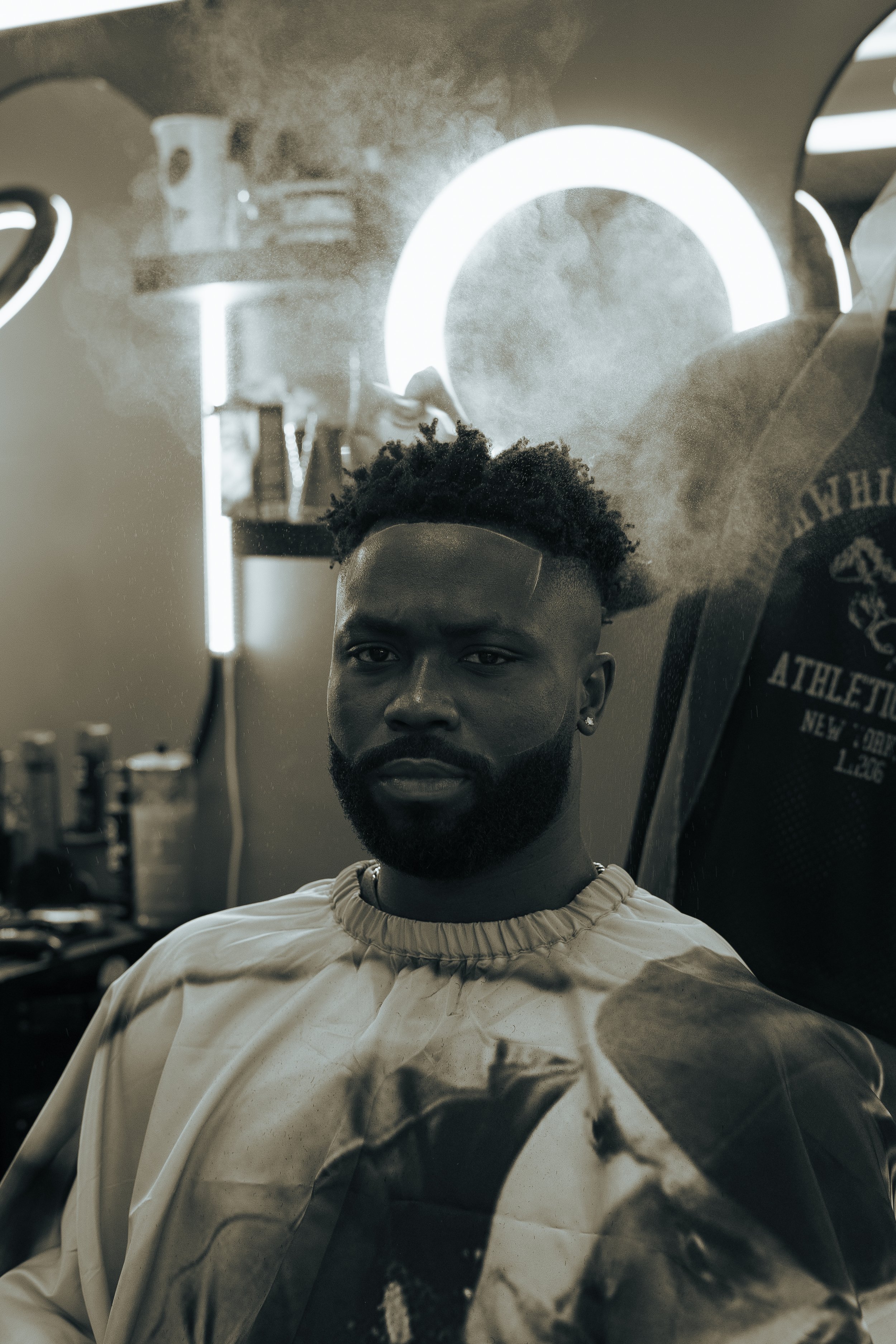 A man with a beard and styled hair is sitting in a barber's chair, wearing a cape, inside a barber shop with neon lighting and several grooming products visible in the background.