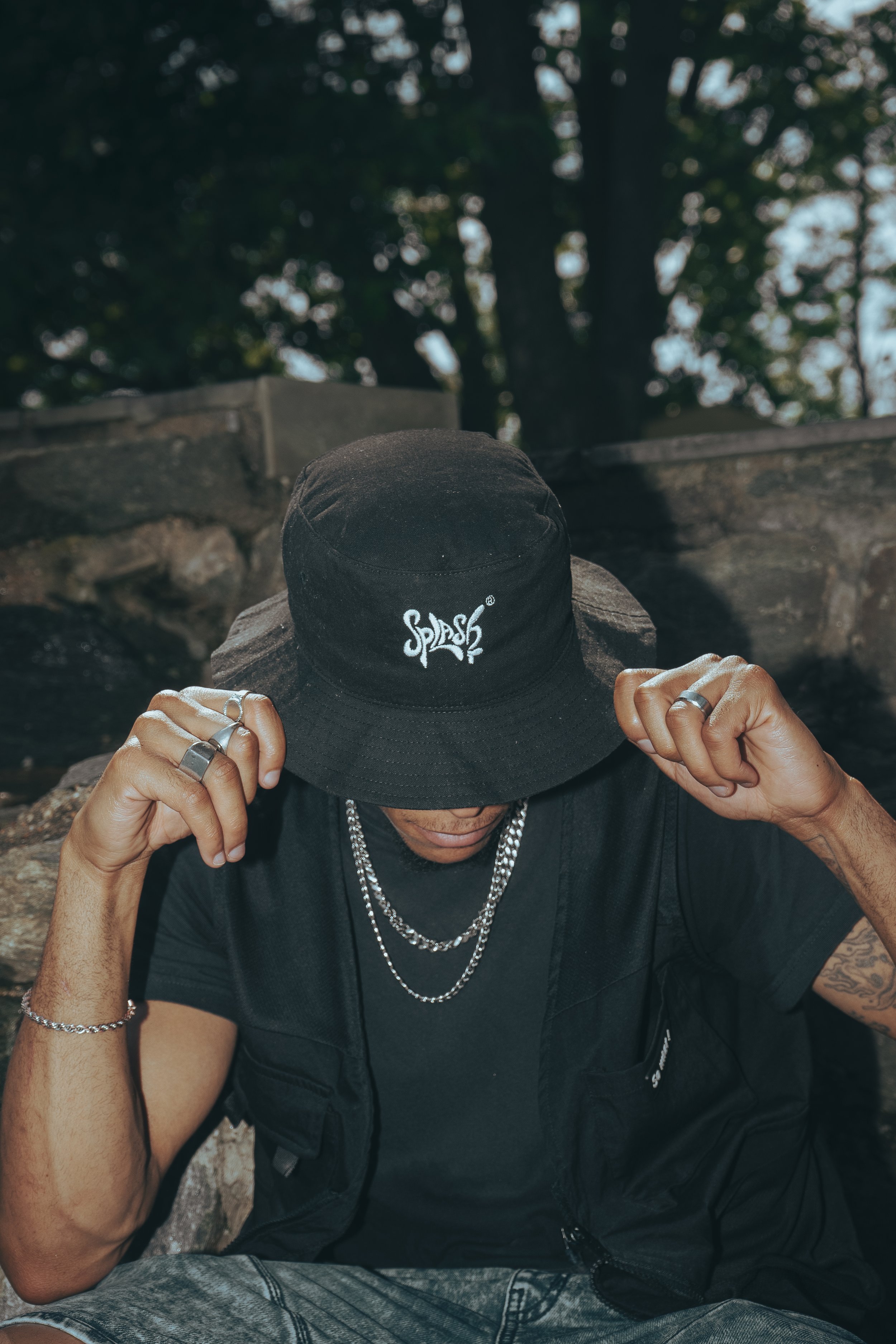 A person wearing a black bucket hat with white writing, multiple silver chains, rings, and a bracelet, adjusting their hat while sitting outdoors near rocks and trees.
