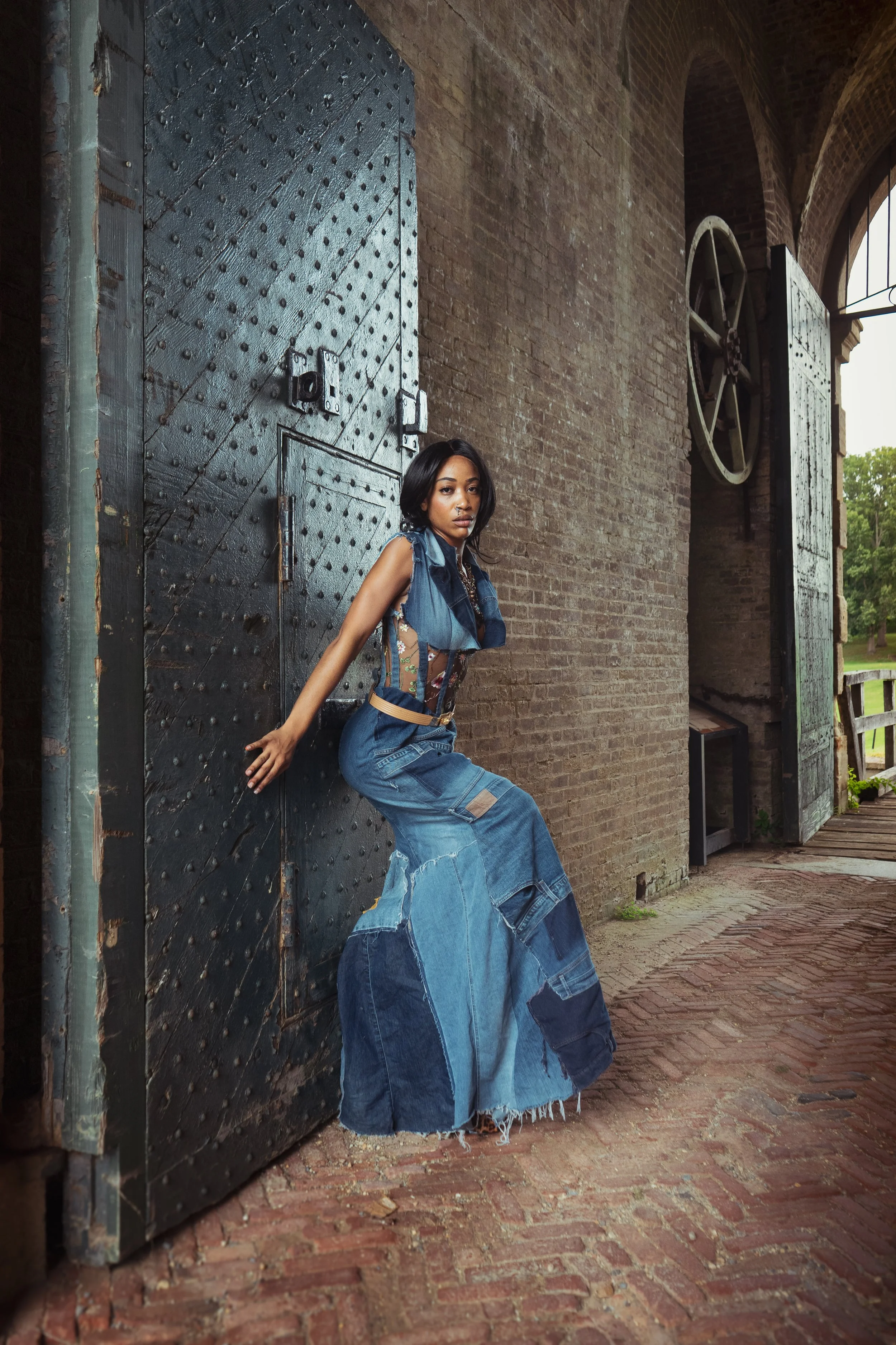 Young woman leaning against a large, black, metal door with rivets, in front of a brick wall with an arched window and large wheel.