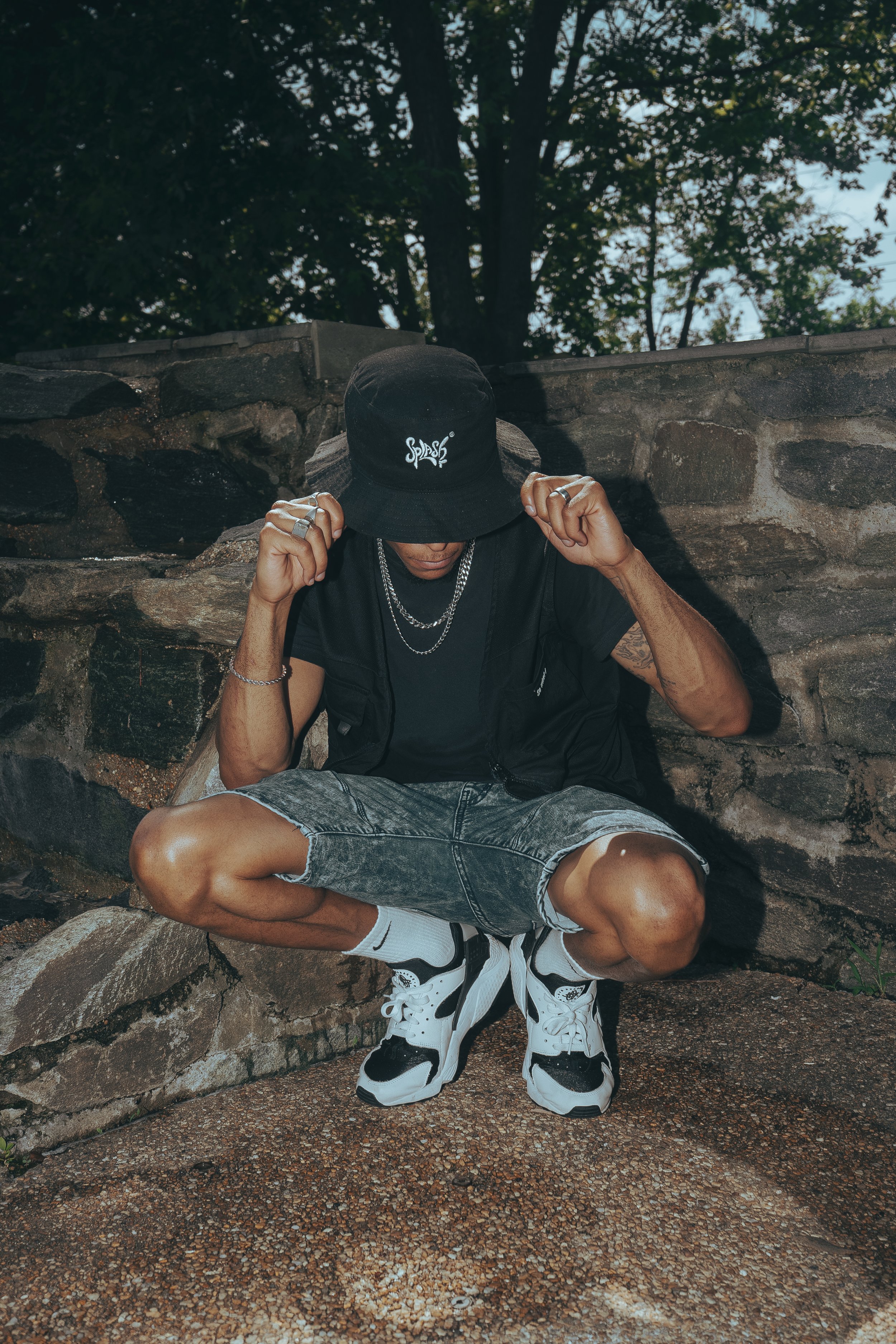 A person dressed in black with a bucket hat, multiple chains, and denim shorts squatting outdoors against a stone wall, adjusting the hat.