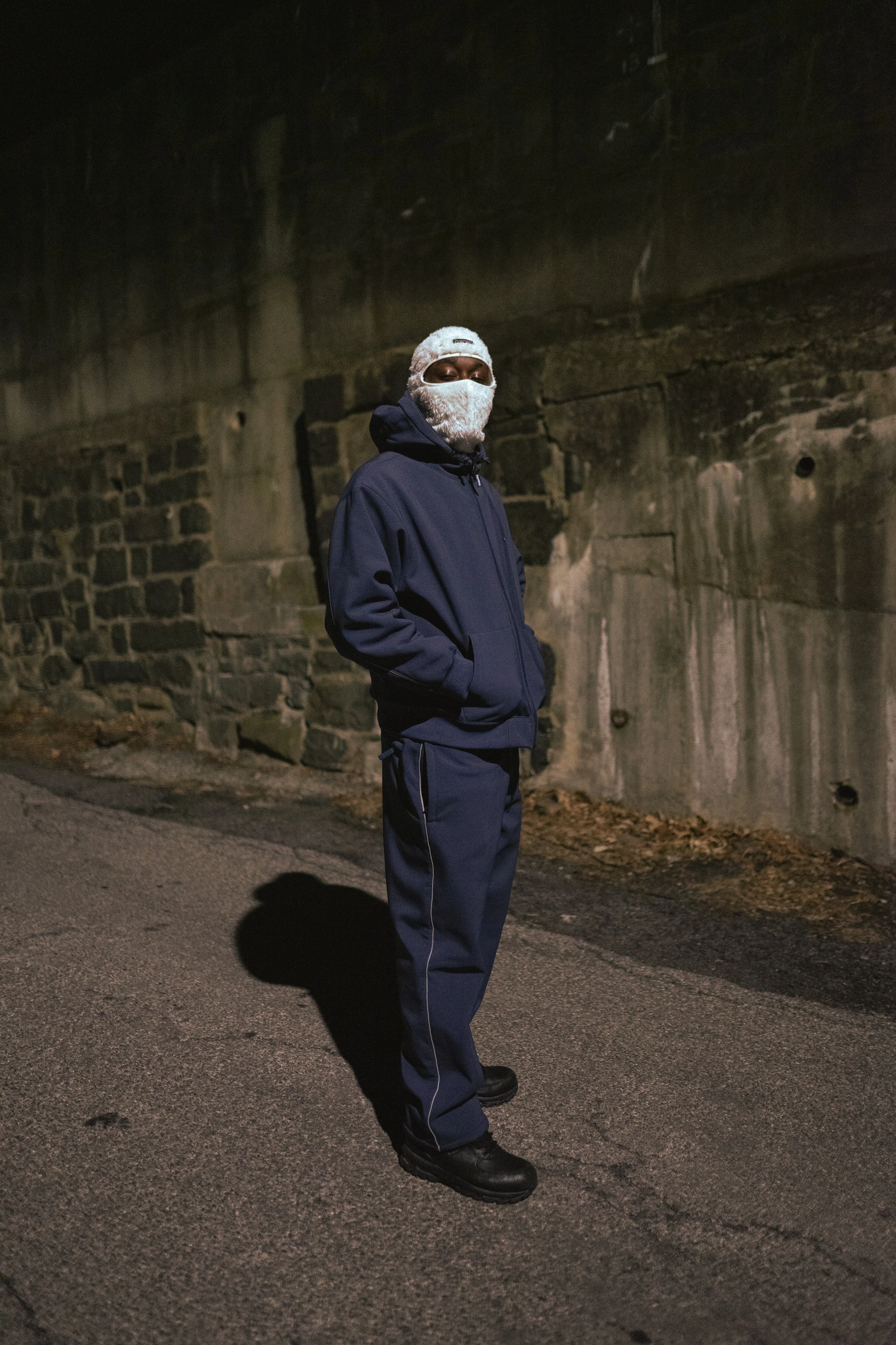 Person in dark clothing and white headscarf, standing on a dimly lit street near a stone wall at night.