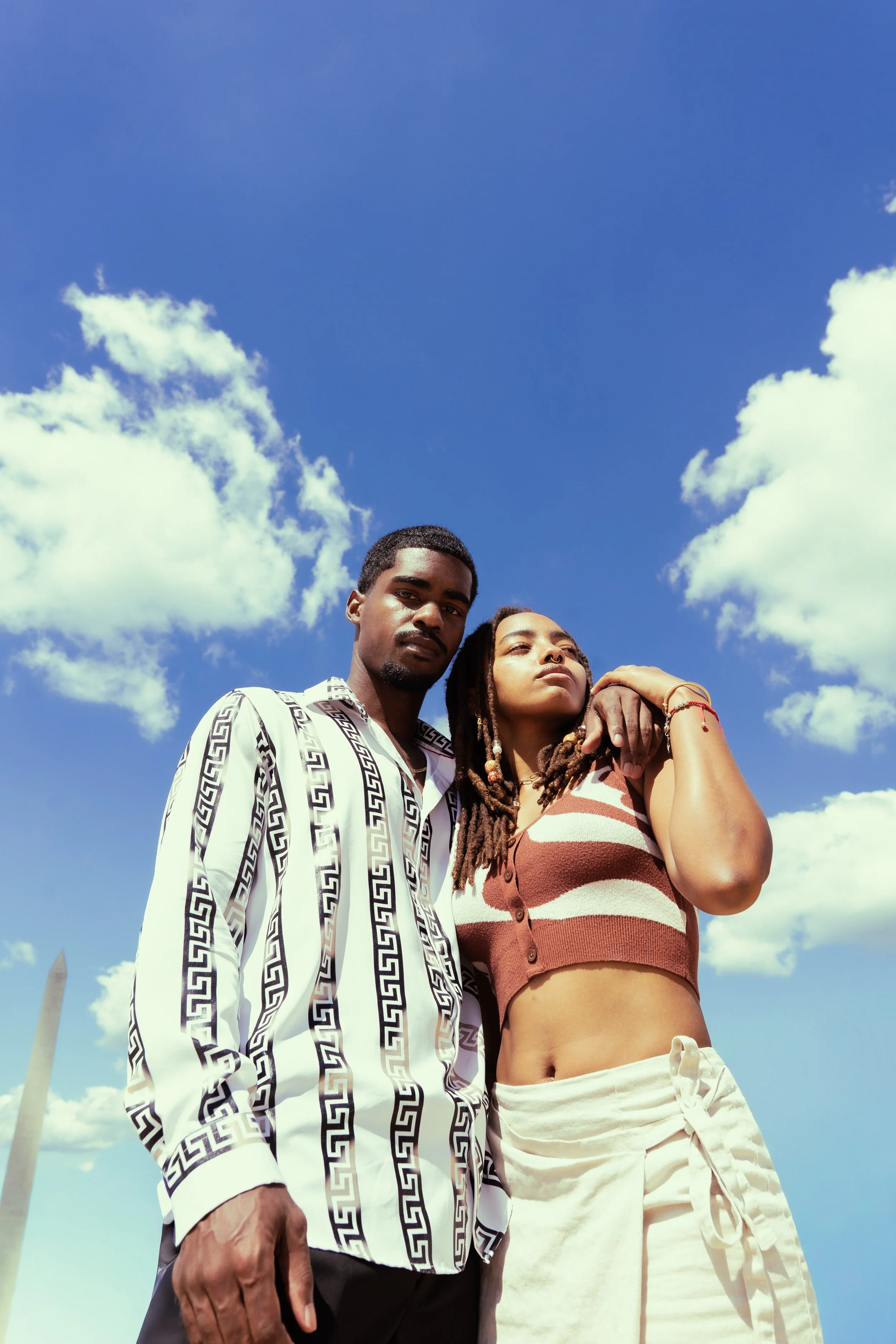 Two young people, a man and a woman, standing closely together outdoors against a blue sky with white clouds. The man has short dark hair and wears a white shirt with black geometric patterns, and the woman has long dreadlocks, and is wearing a sleev