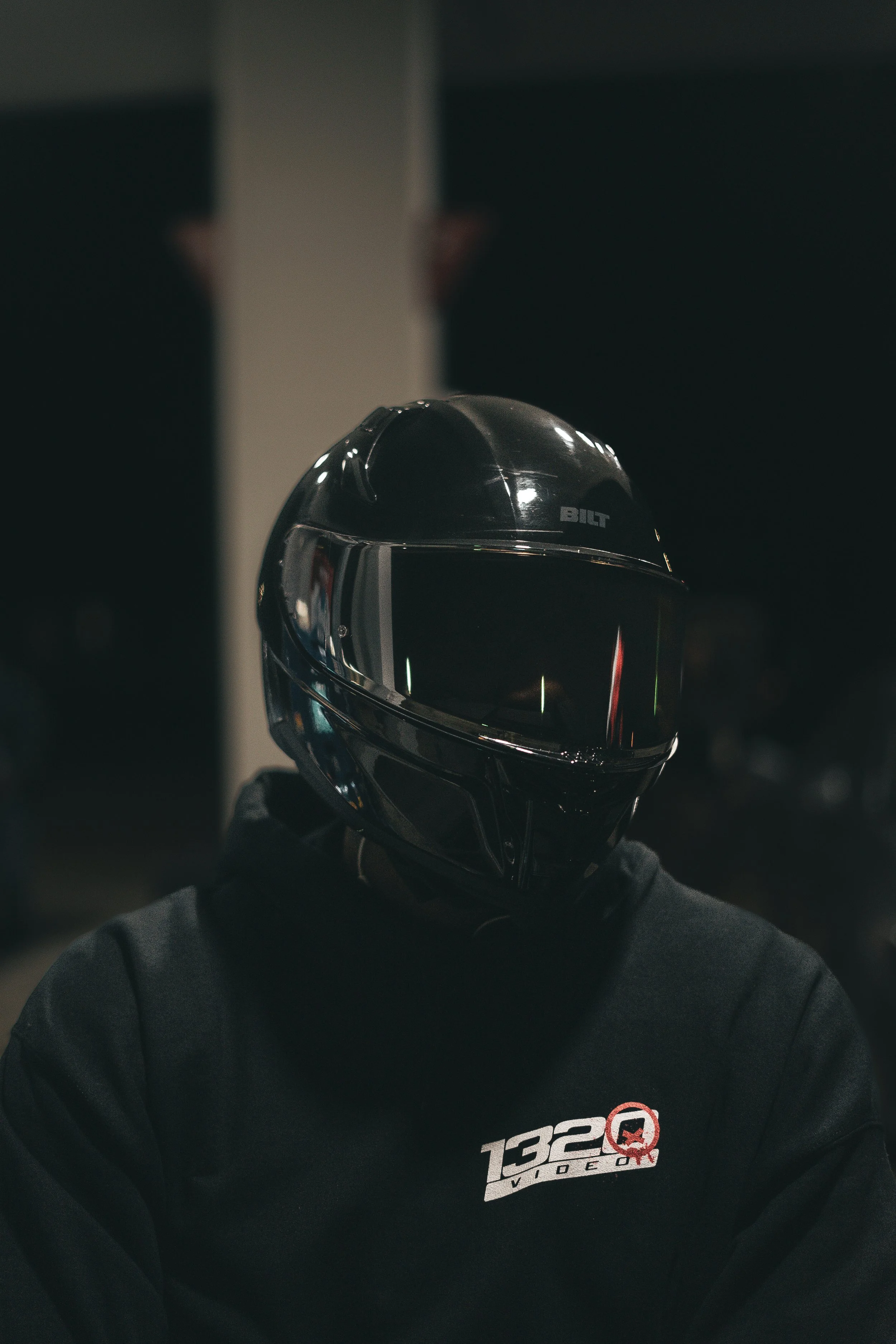 Person wearing a black motorcycle helmet and dark hoodie with "1320 Video" logo, in a dimly lit setting.