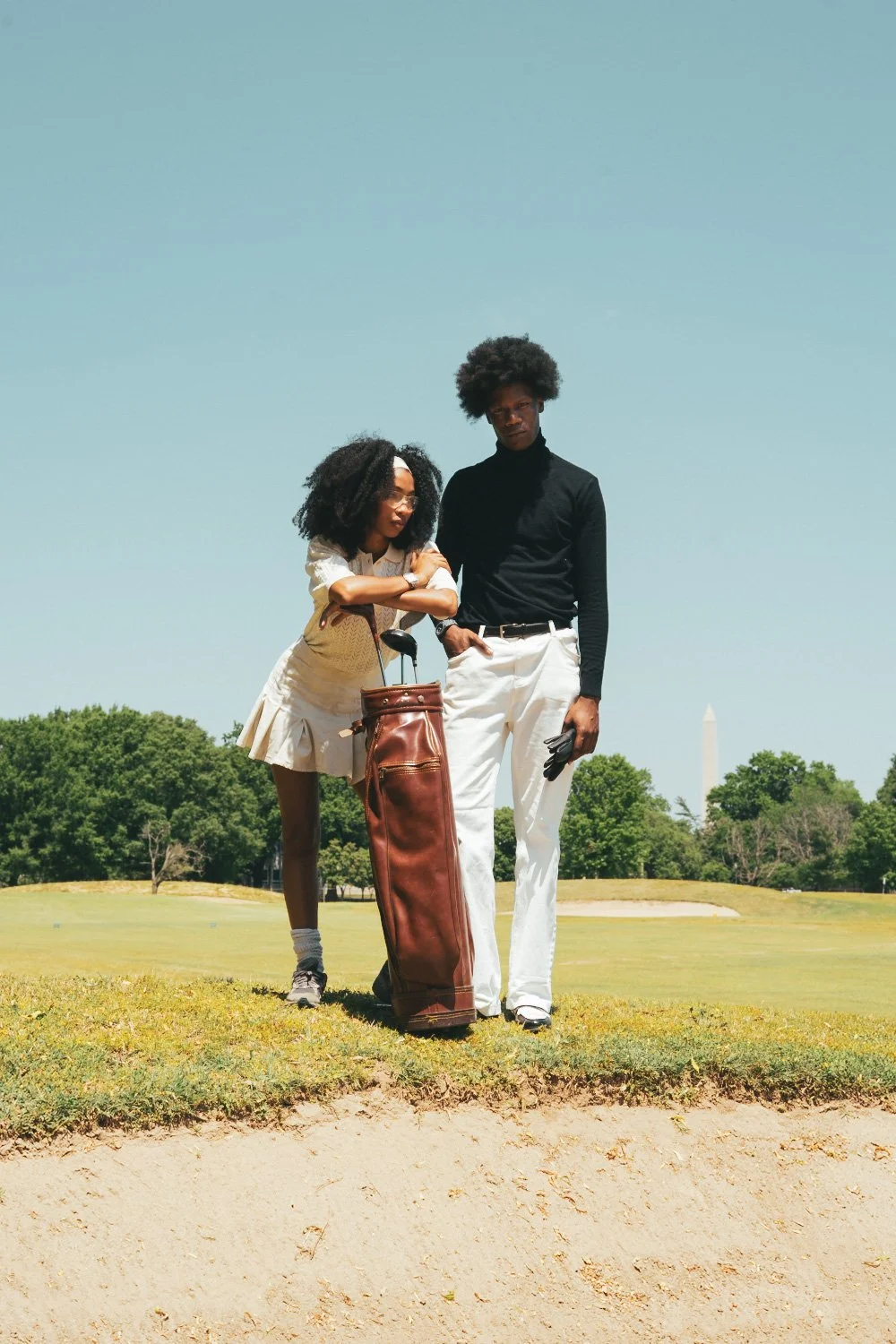 Two people, a woman and a man, stand on a golf course with a flag and trees in the background. The woman leans on a golf bag, wearing a cream-colored outfit and sunglasses. The man is dressed in a black turtleneck and white pants, holding a golf glov