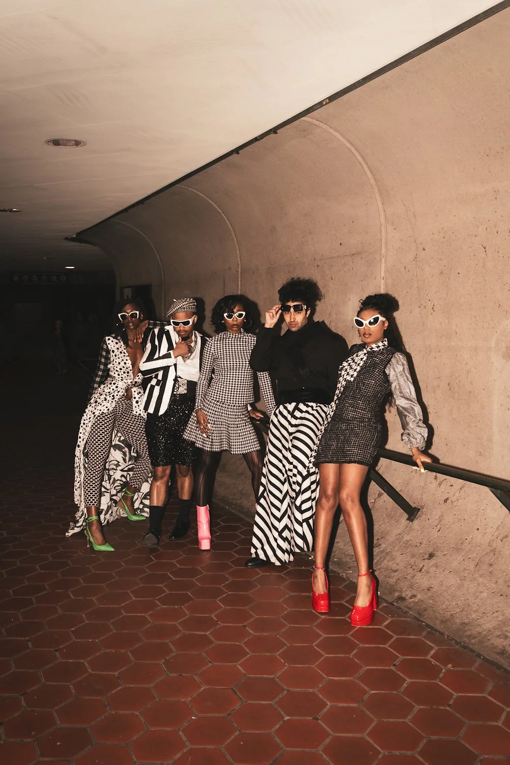 Group of six stylish people wearing black and white outfits with bold patterns, sunglasses, and high heels, posing in a dimly lit indoor space with a curved concrete wall and tiled floor.
