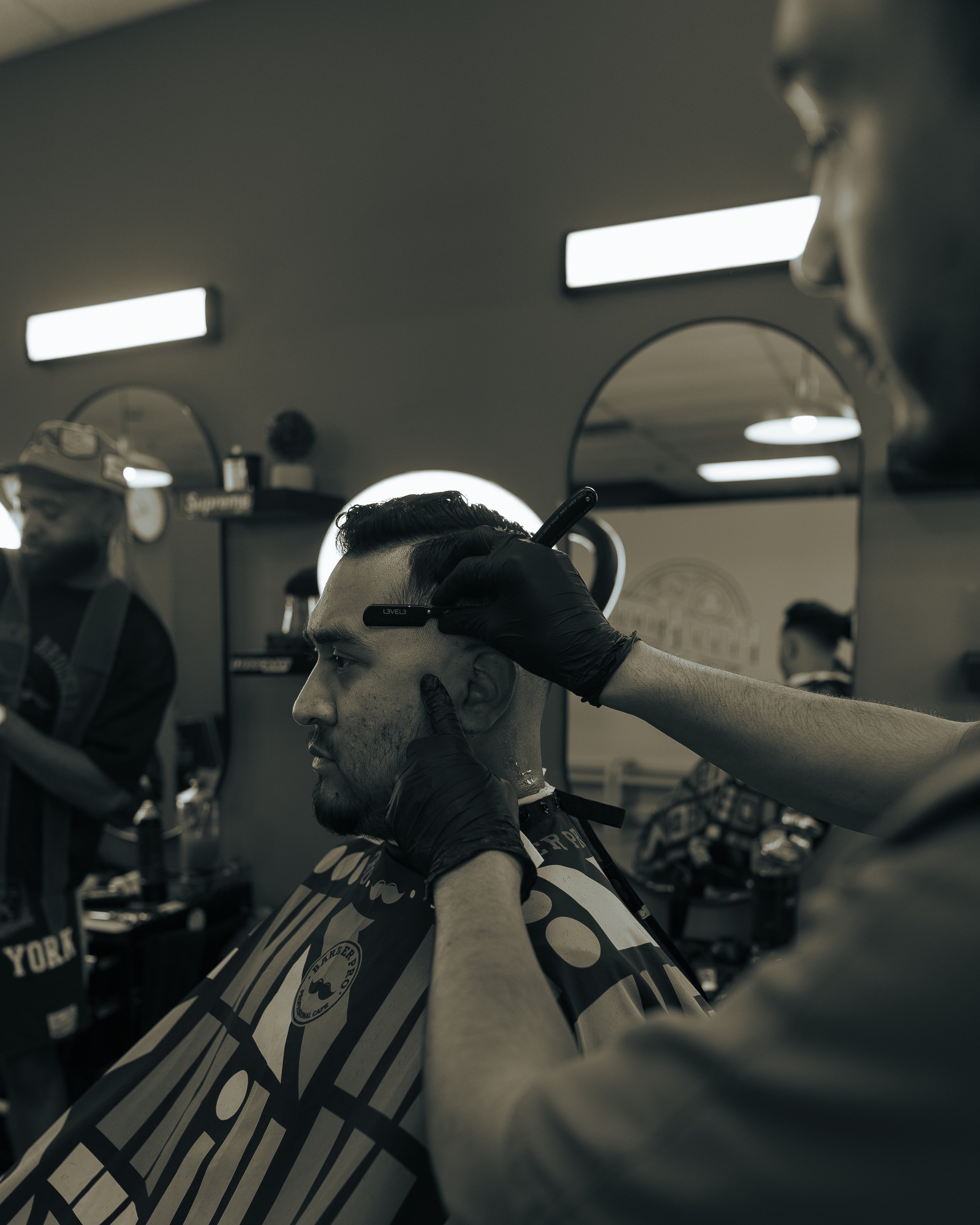 A man getting a haircut in a barbershop, with a barber trimming his hair using clippers, while wearing black gloves.