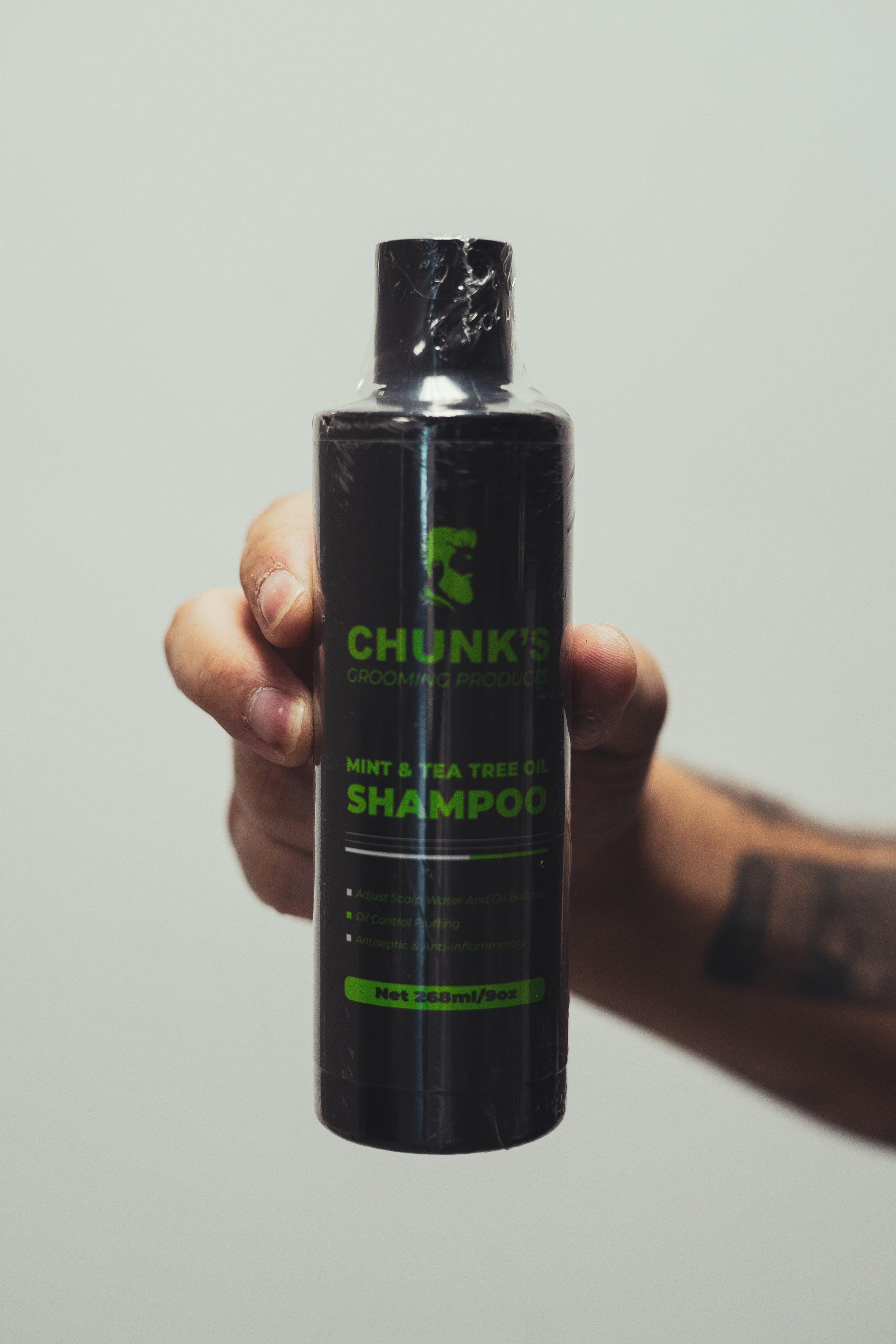 A person holding a black bottle of Chunk's Grooming Products Mint & Tea Tree Oil Shampoo against a plain white background.