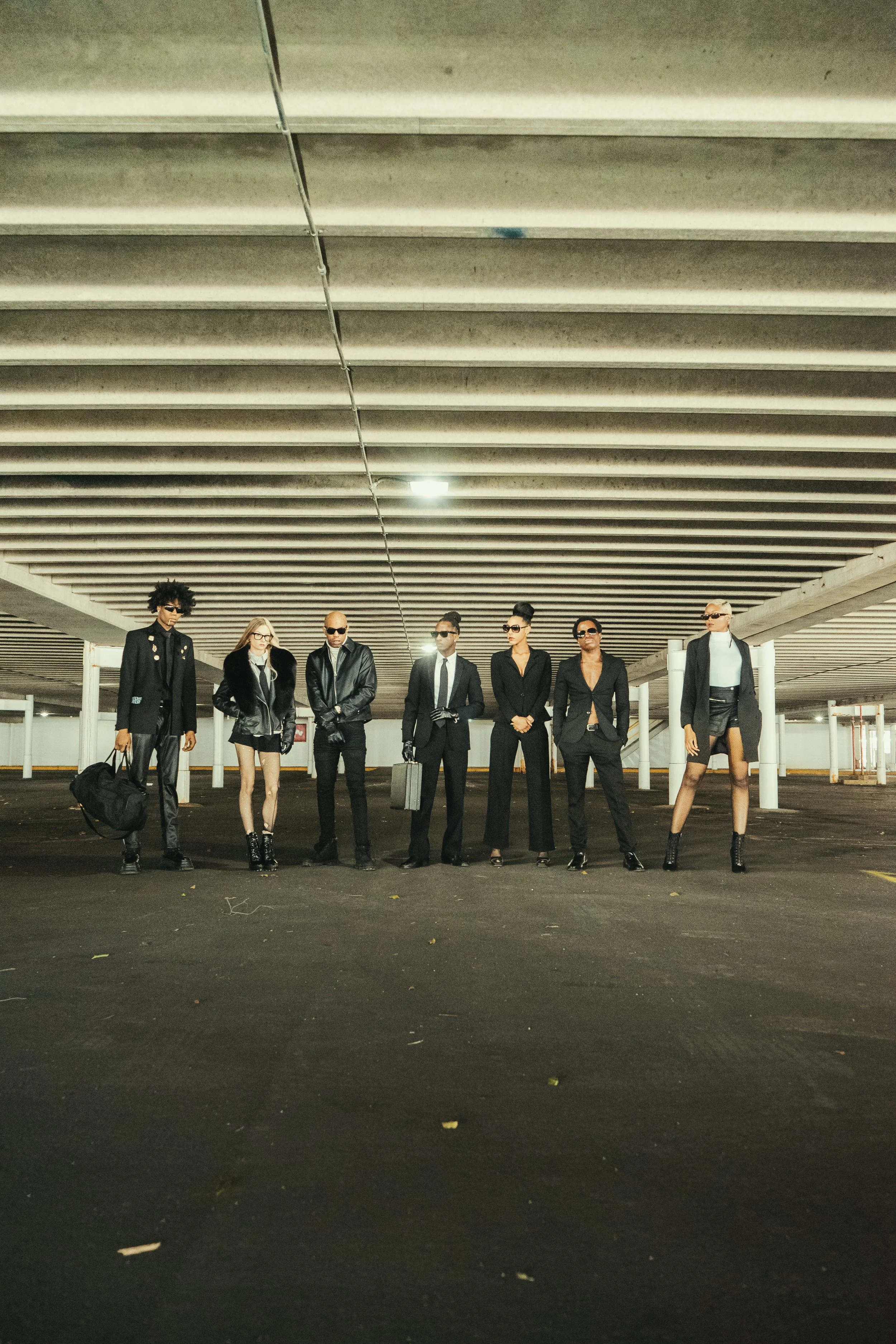 A group of seven fashionable individuals posing under a modern parking garage ceiling. They are dressed in stylish black clothing, with some wearing sunglasses and carrying bags or briefcases.