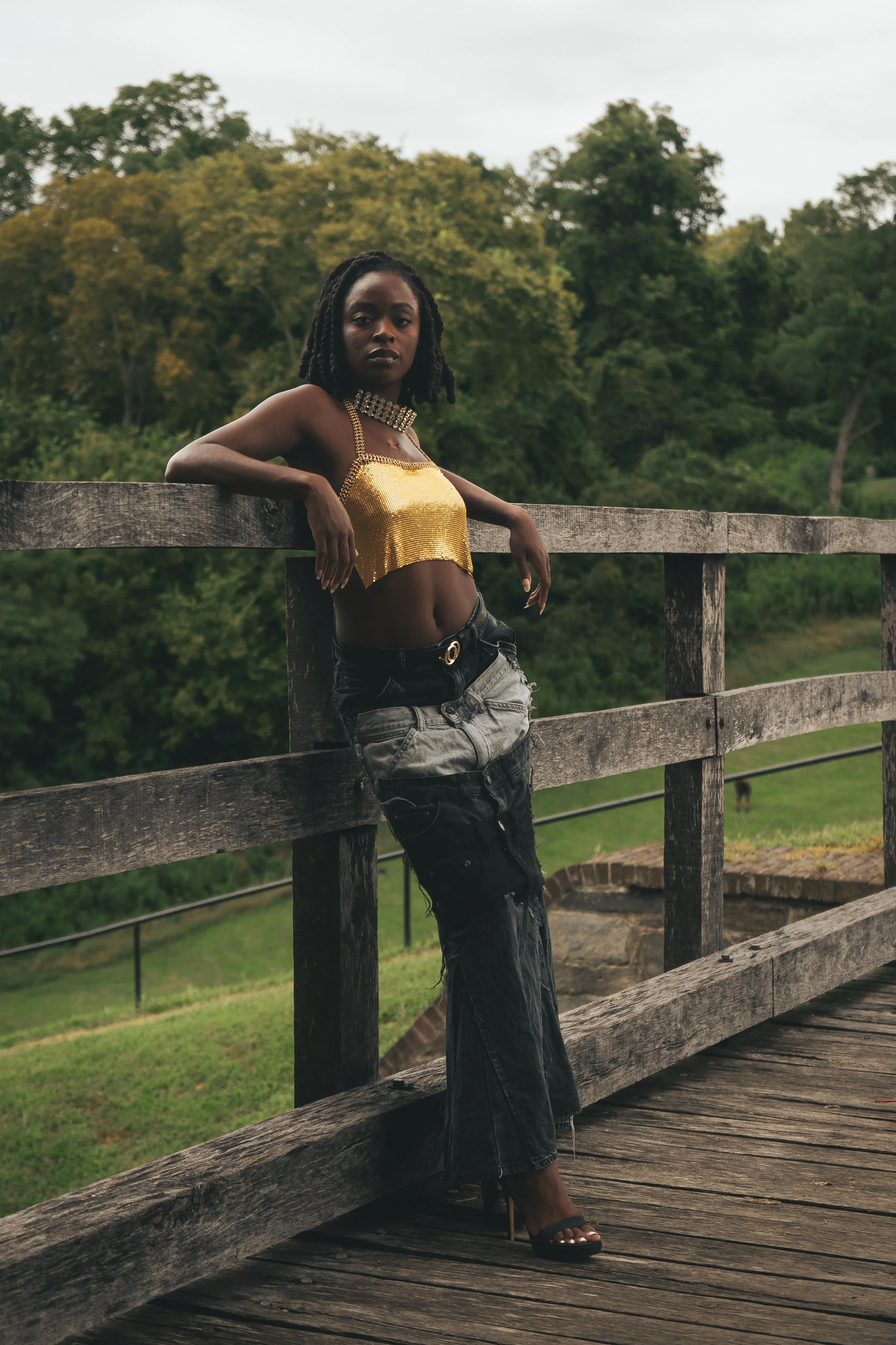 A woman with dark skin and medium-length dreadlocks poses on a wooden bridge with a scenic forested background, wearing a gold crop top, black distressed jeans, and black high-heeled sandals.