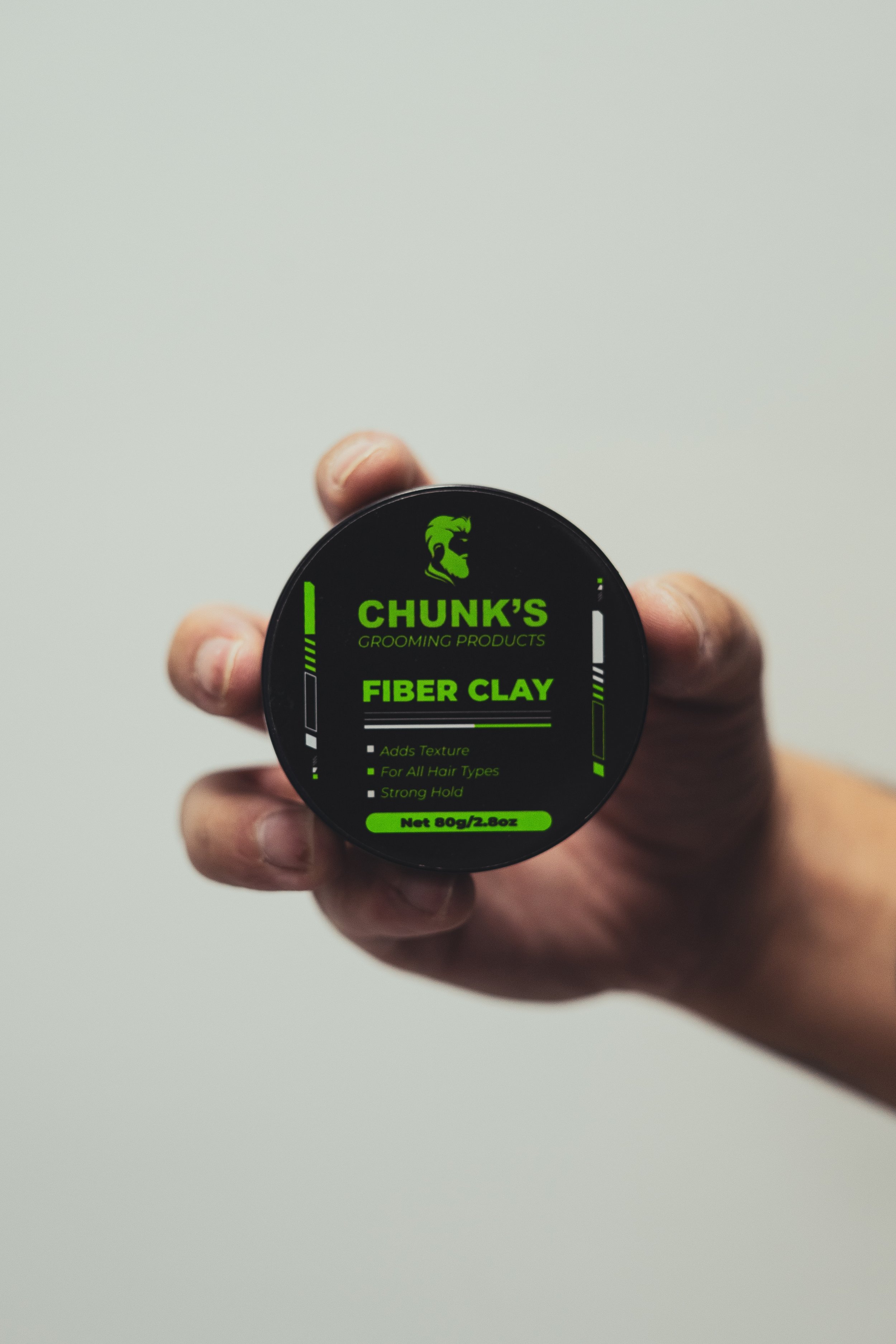 A hand holding a round container of Chunk's Grooming Products Fiber Clay with a black and neon green label.