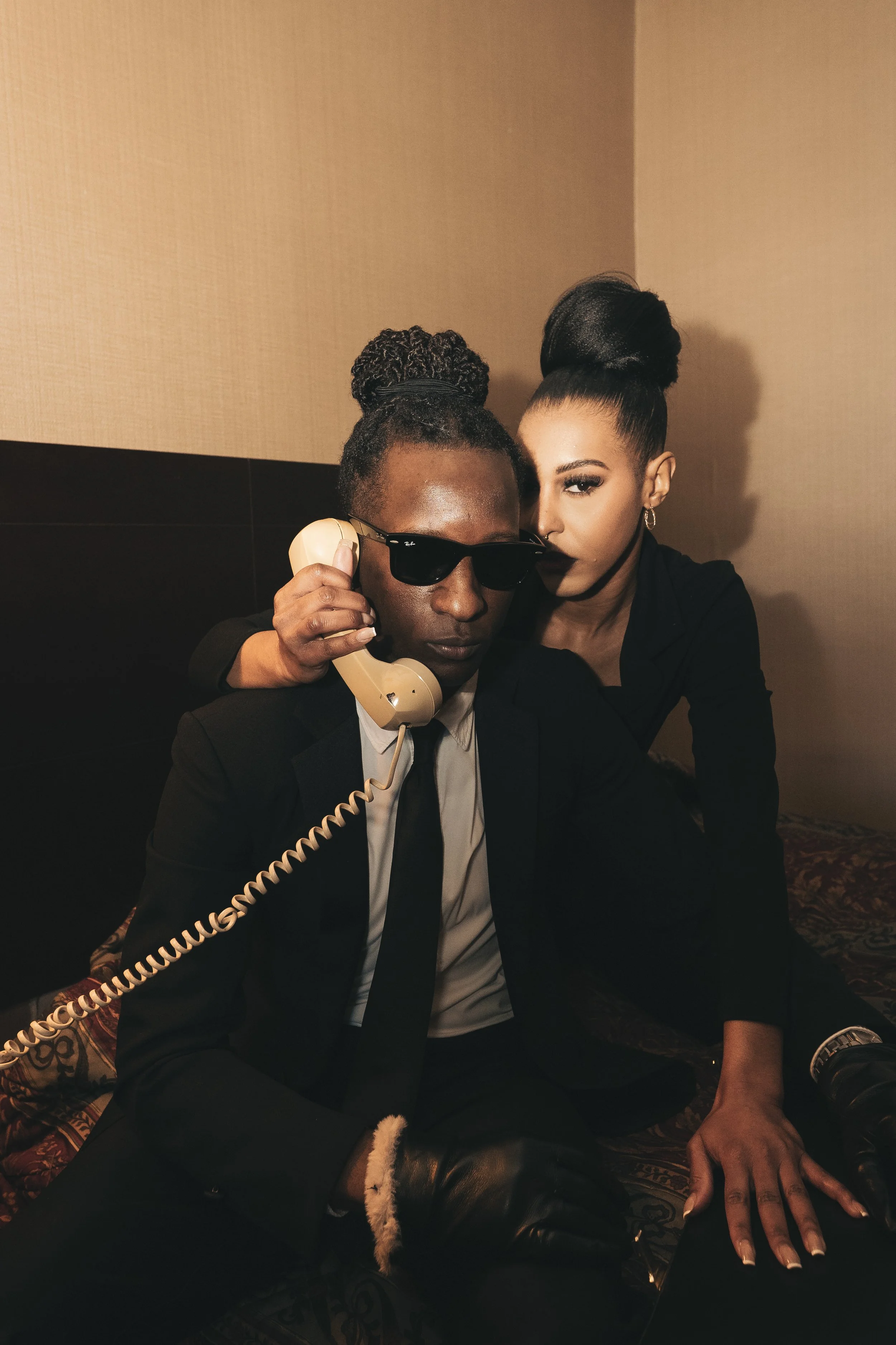 A man in a black suit and sunglasses holding a vintage telephone receiver next to a woman with styled hair and makeup, dressed in black, with her arm around his shoulder, indoor setting.