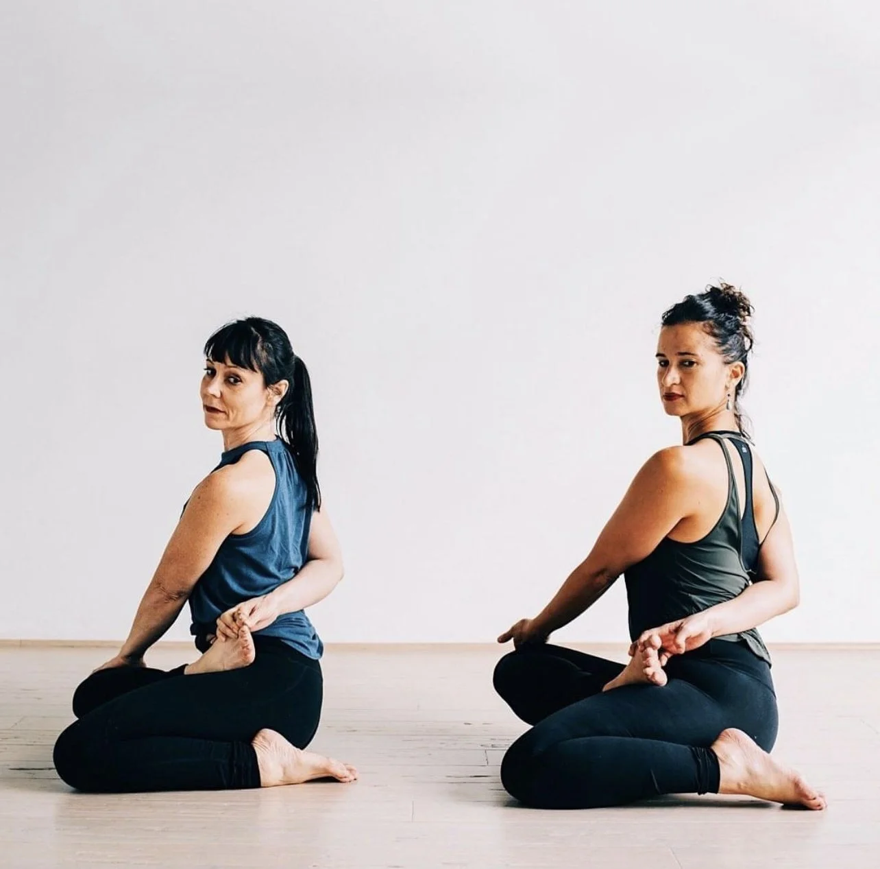 Two women practicing yoga while sitting cross-legged on the floor, holding their feet with their hands, against a plain white wall.