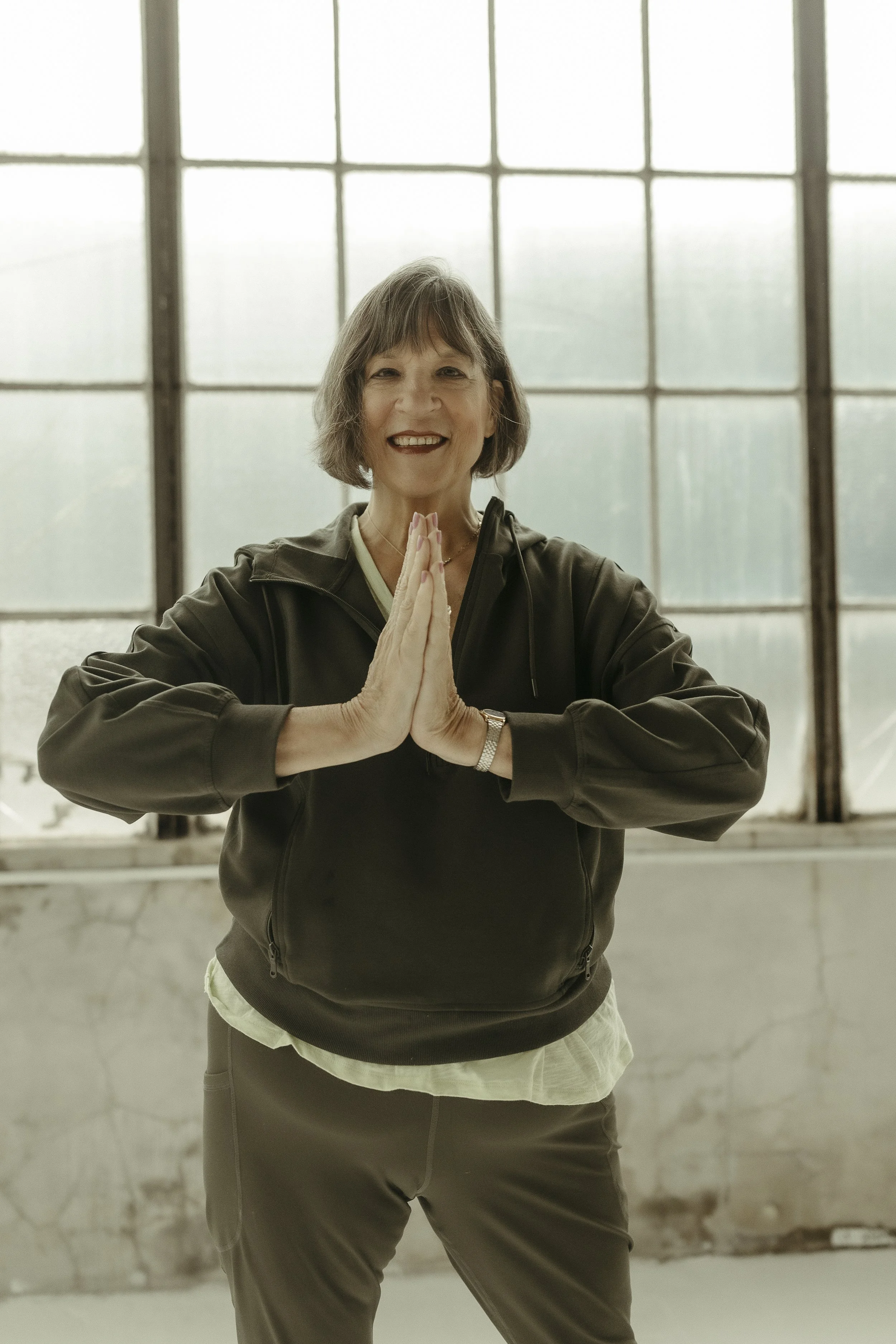 Two men practicing yoga indoors; one man with tattoos assisting the other in a yoga pose on a black mat in front of large industrial-style windows.