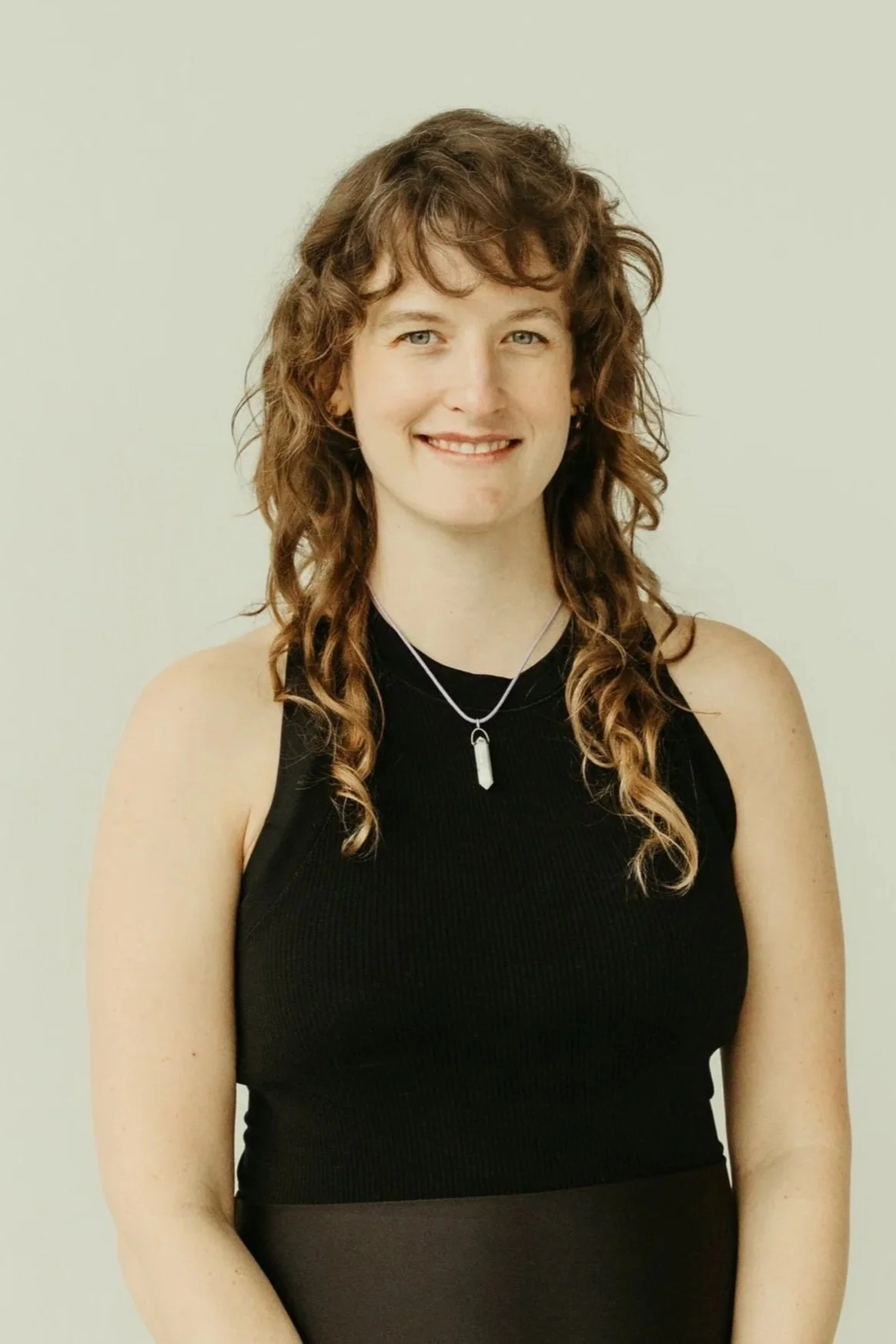 A woman sitting against a plain light background, wearing a black sleeveless top and black shorts, smiling and looking at the camera.