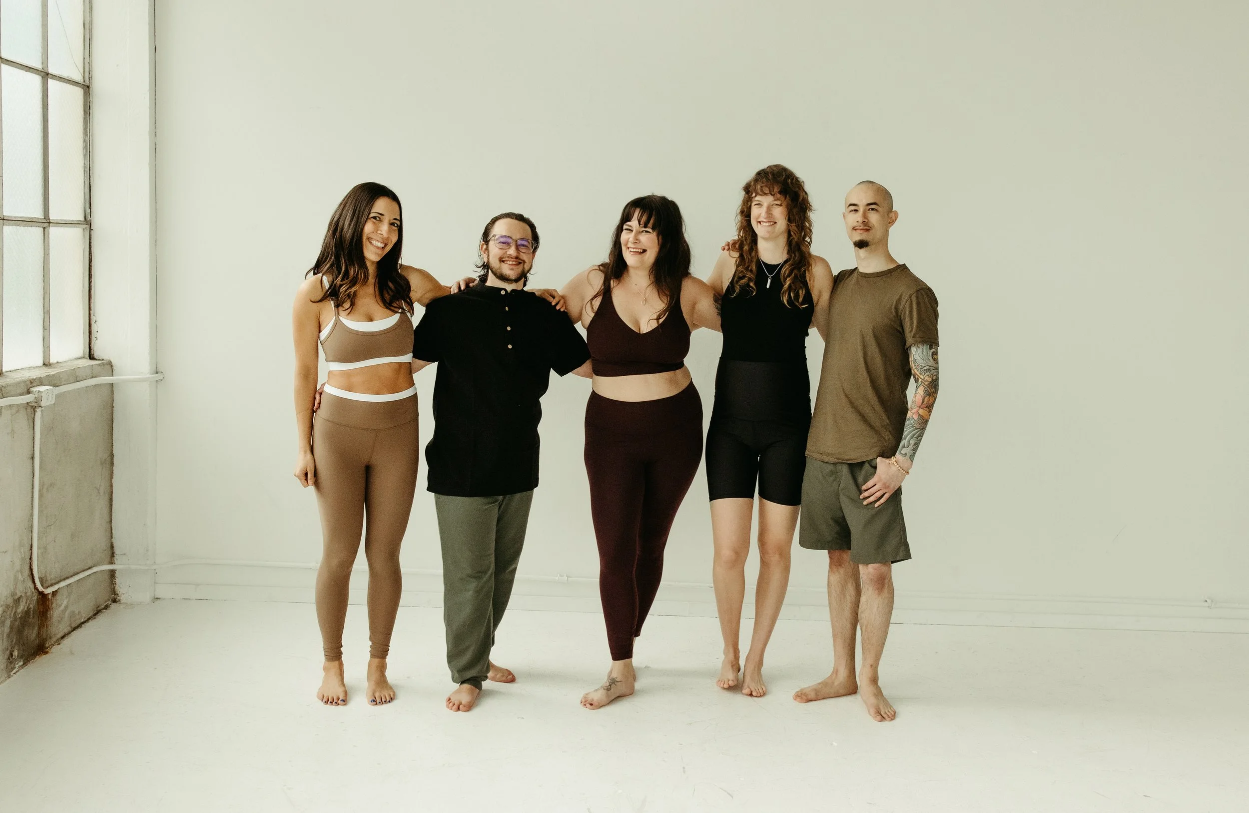 Group of five diverse people smiling and standing together indoors, with their arms around each other's shoulders, in a bright studio setting.