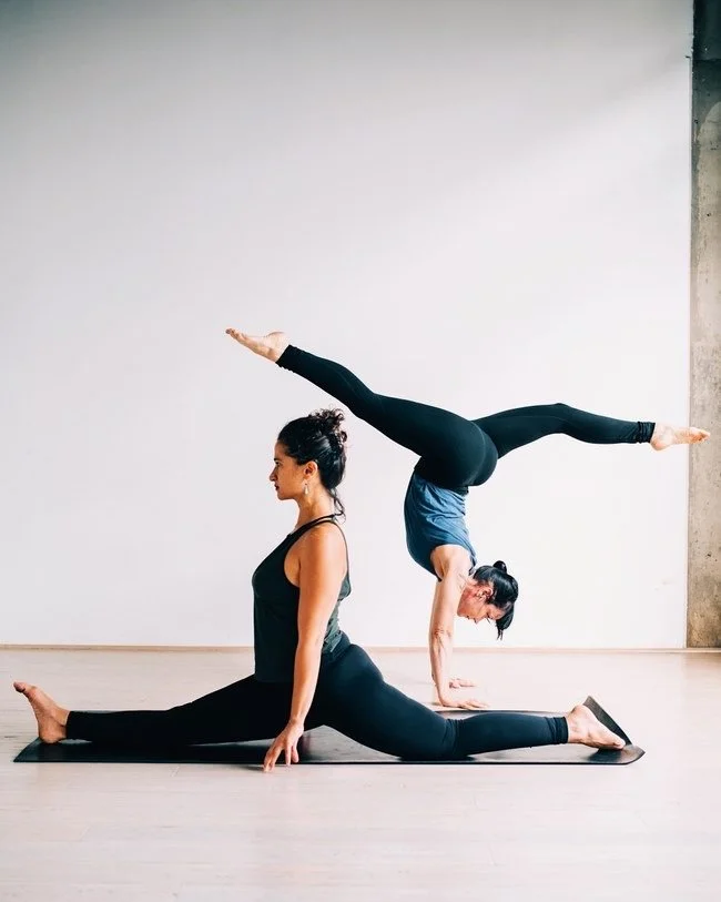 Two women practicing yoga indoors; one is in a split pose on the floor with her legs extended and hands on the ground, while the other is balancing in a handstand on her partner's back.