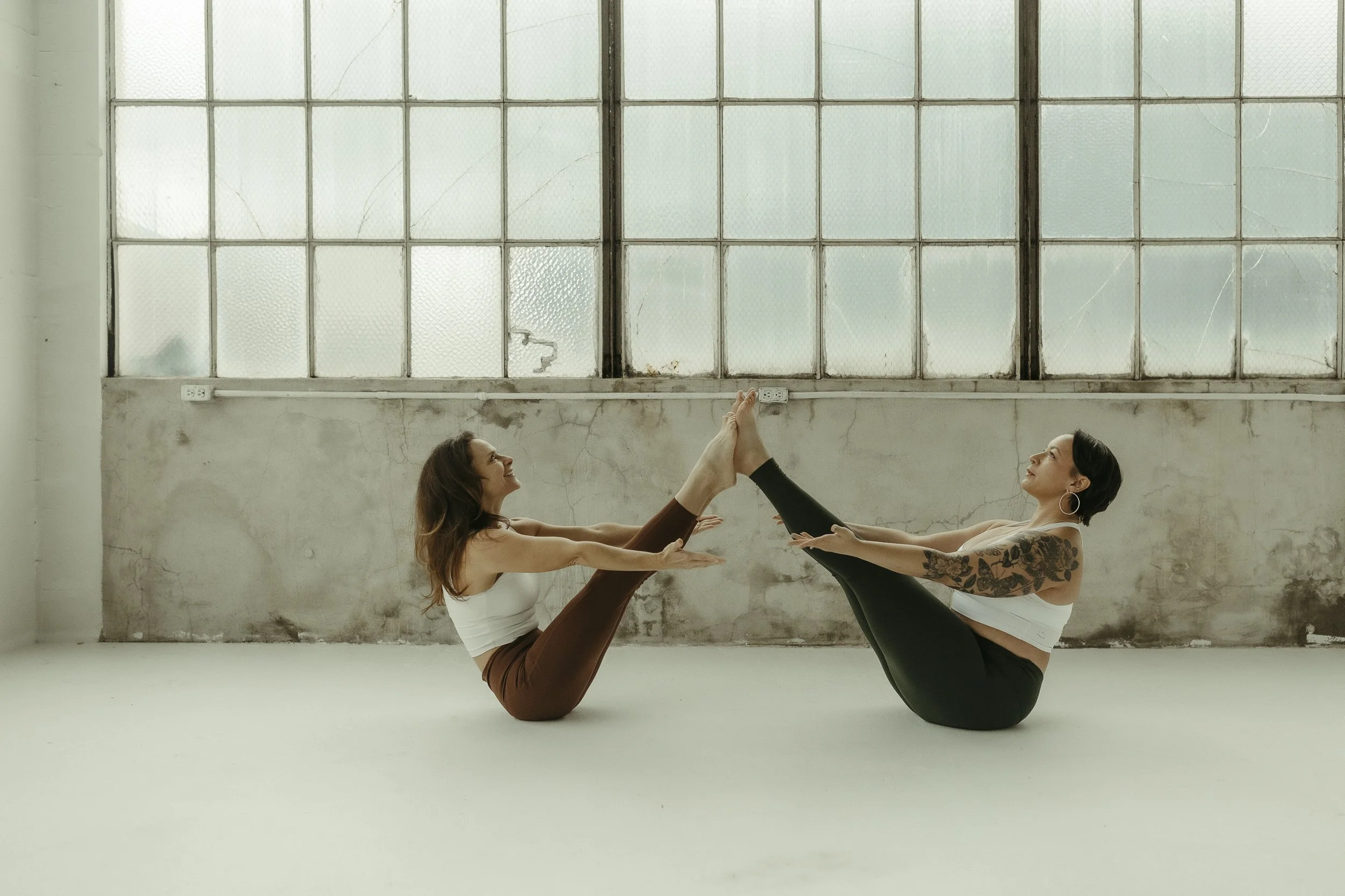 Two women practicing acro yoga indoors, one in a plank position on the floor, the other standing on her partner's back with her arm raised, both smiling.