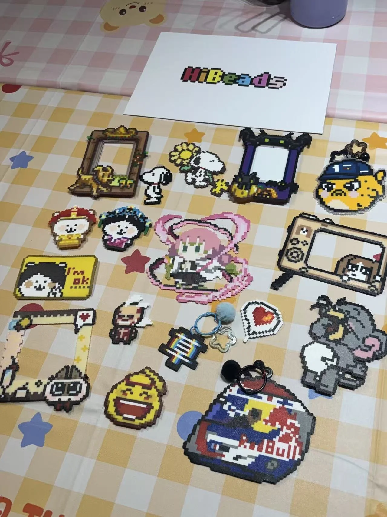 A collection of pixel art characters and items displayed on a table with a checkered tablecloth. The items include various cute characters, a mirror, a photo frame, a Red Bull can, and other small decorative pieces.