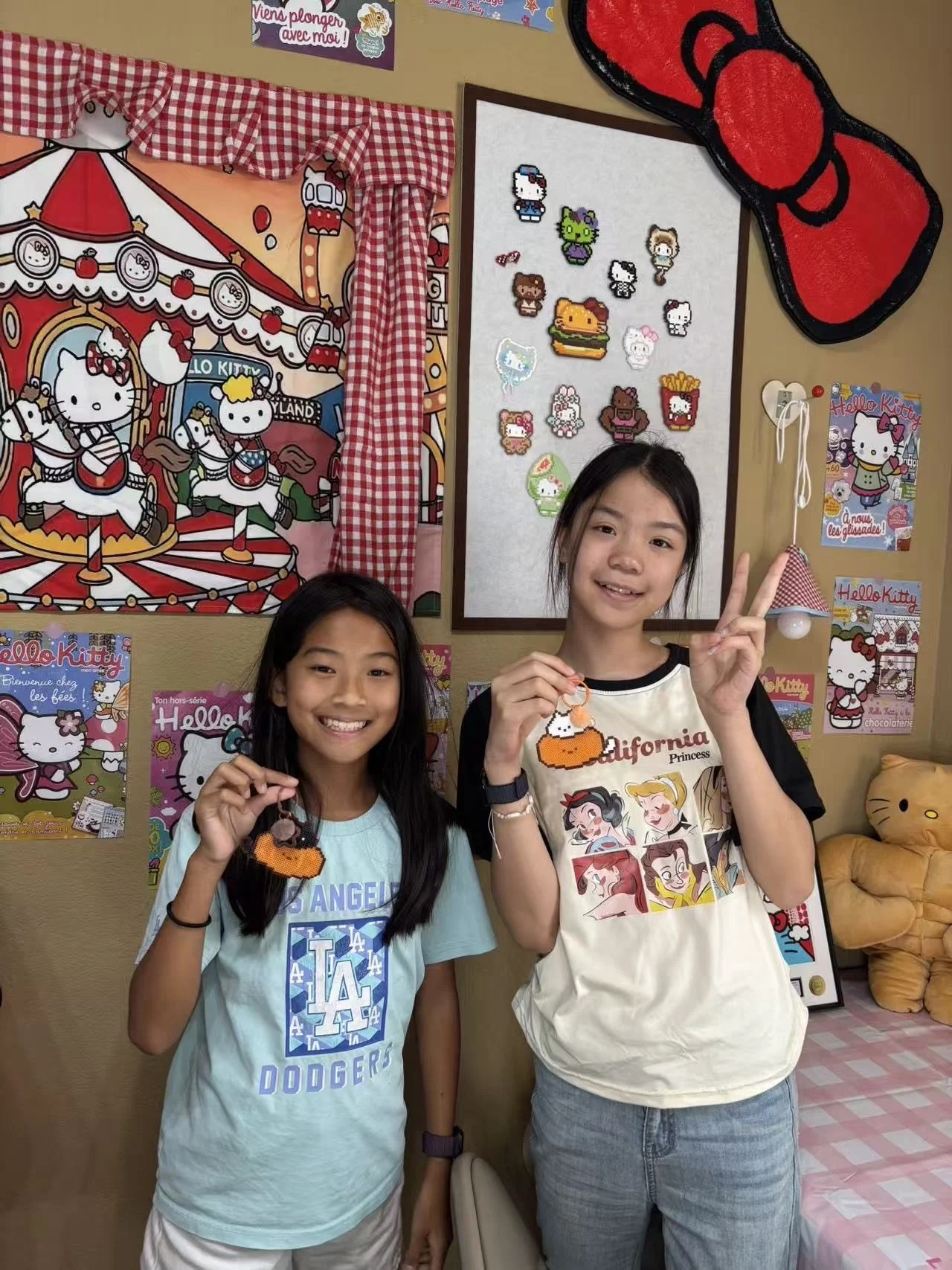 Two girls smiling and holding Hello Kitty plush keychains, standing in front of a wall decorated with Hello Kitty posters and plush toys, with a Hello Kitty carousel illustration on the wall behind them.