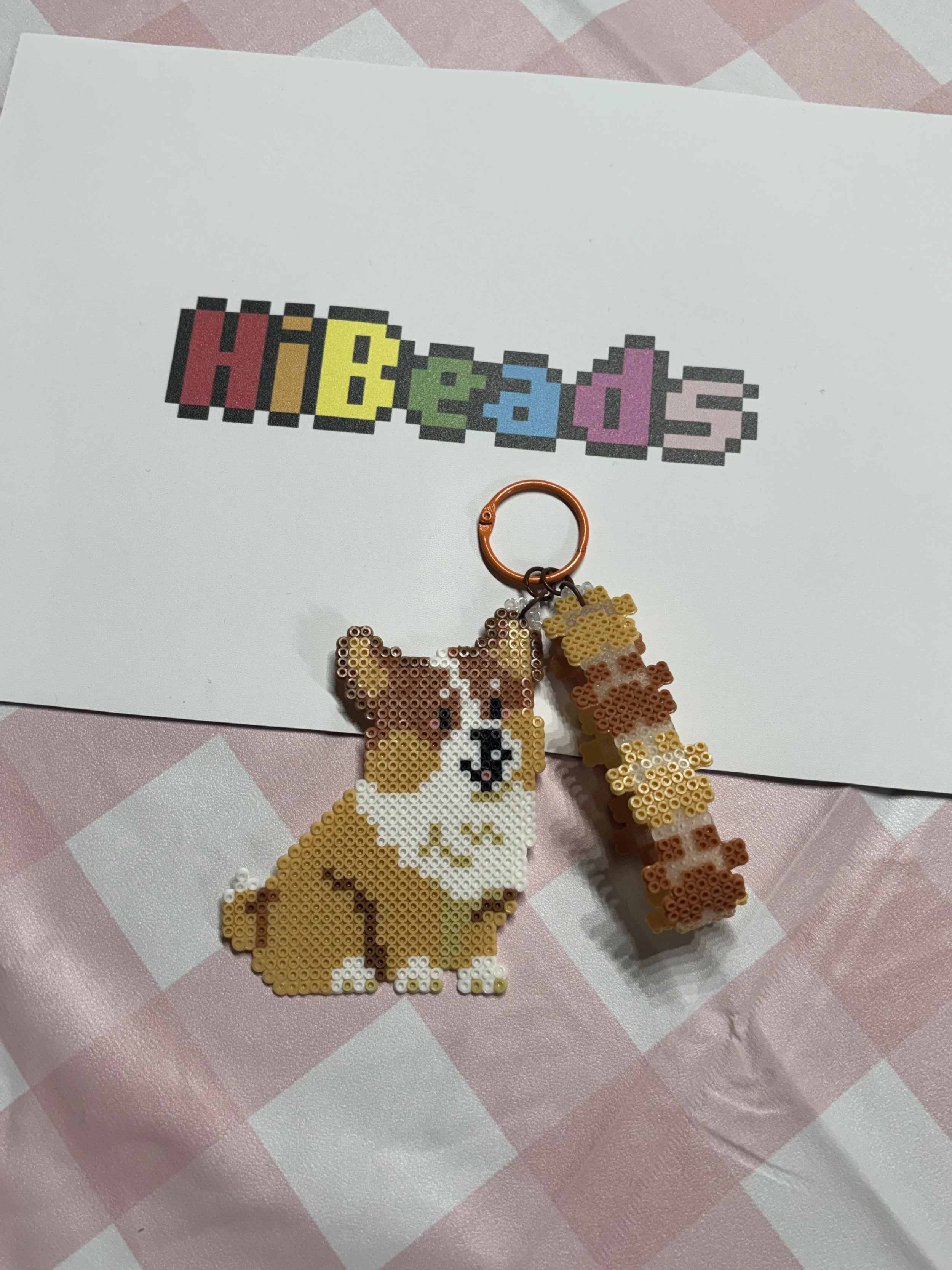 A pixel art-style card with the word 'HIBEADS' in colorful block letters, a dog-shaped cross-stitch keychain, and a small bone-shaped beadwork keychain, all placed on a pink and white checkered tablecloth.