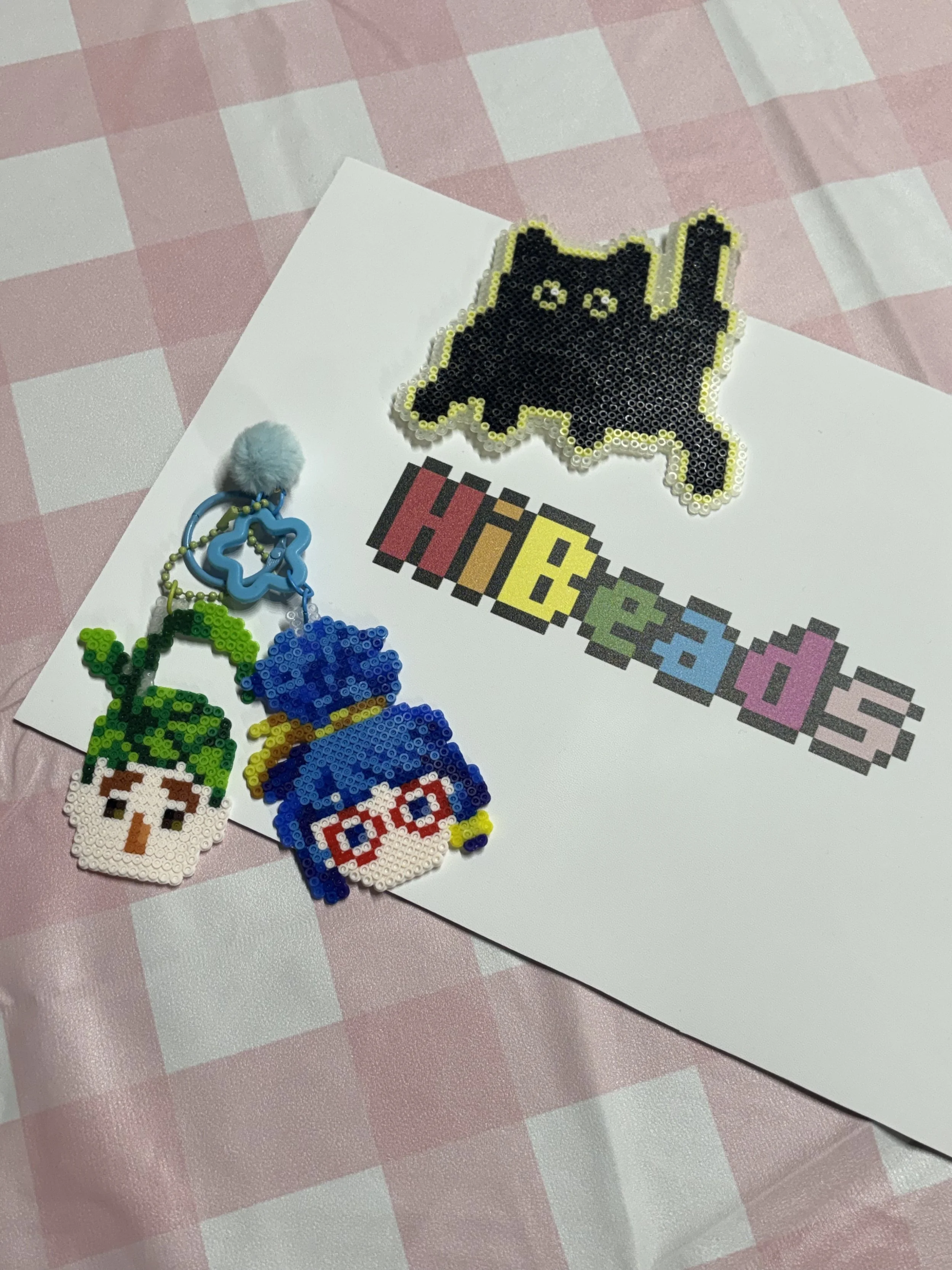 Beaded keychains featuring a character with green hair, a character with glasses and blue hair, and a fluffy gray pom-pom, all placed on a white paper with a pixelated 'hibeads' text, on a pink and white checkered tablecloth.