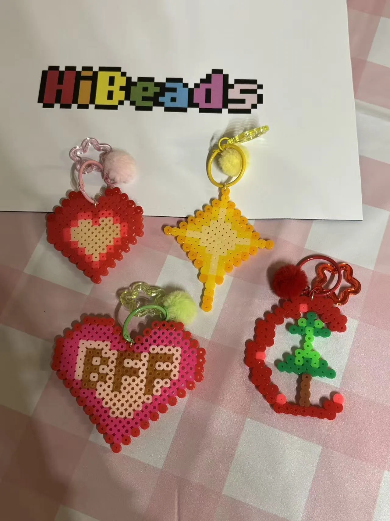 Four pixel art-style beaded keychains with pom-poms, shaped as a heart, a star, a cherry, and a Christmas tree, displayed on a pink and white checkered surface with a white paper in the background reading "HIBeads" in colorful pixelated letters.